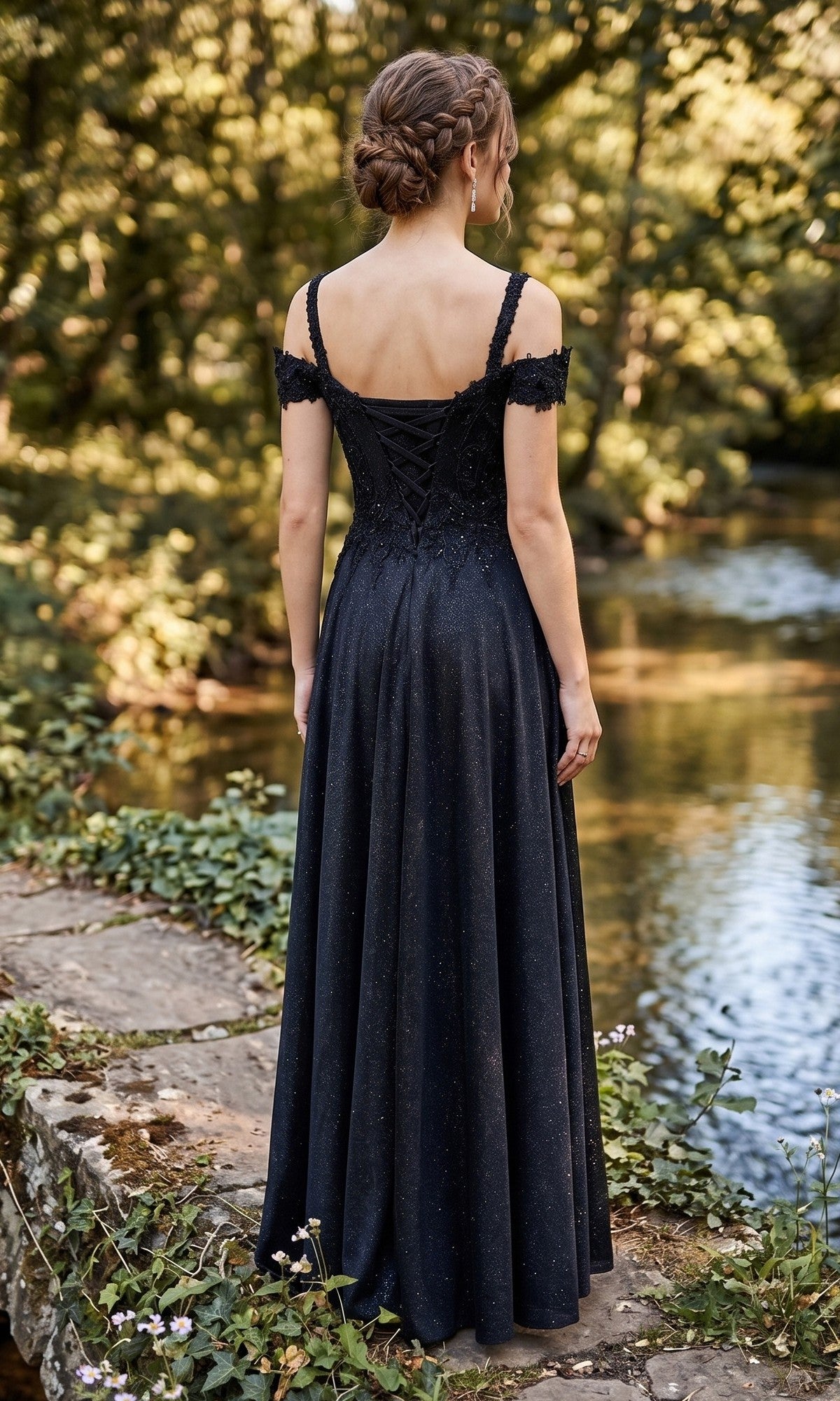 A woman with braided hair stands on a stone path beside a river, surrounded by greenery, wearing the Chicas J169 Long Prom Dress—an elegant black off-shoulder gown with an embellished bodice.