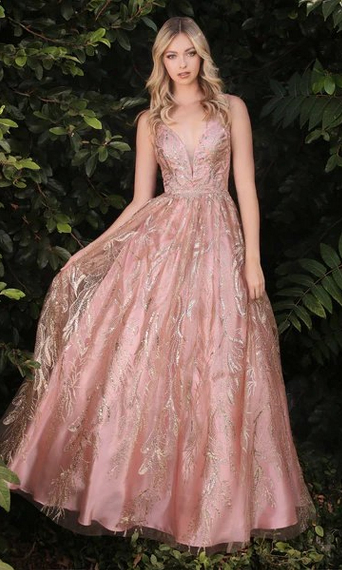 A woman stands outdoors in front of greenery, wearing the Sleeveless Glitter-Print Long Prom Dress J812, a floor-length gown with pink metallic leaf patterns and a deep V-neckline.