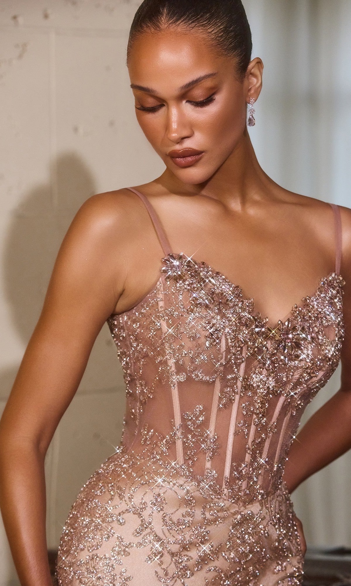 A woman stands indoors, looking down in the Ladivine J889 Long Prom Dress—an elegant nude gown featuring a fitted silhouette, sheer panels, a corset bodice, and intricate beaded floral embellishments.