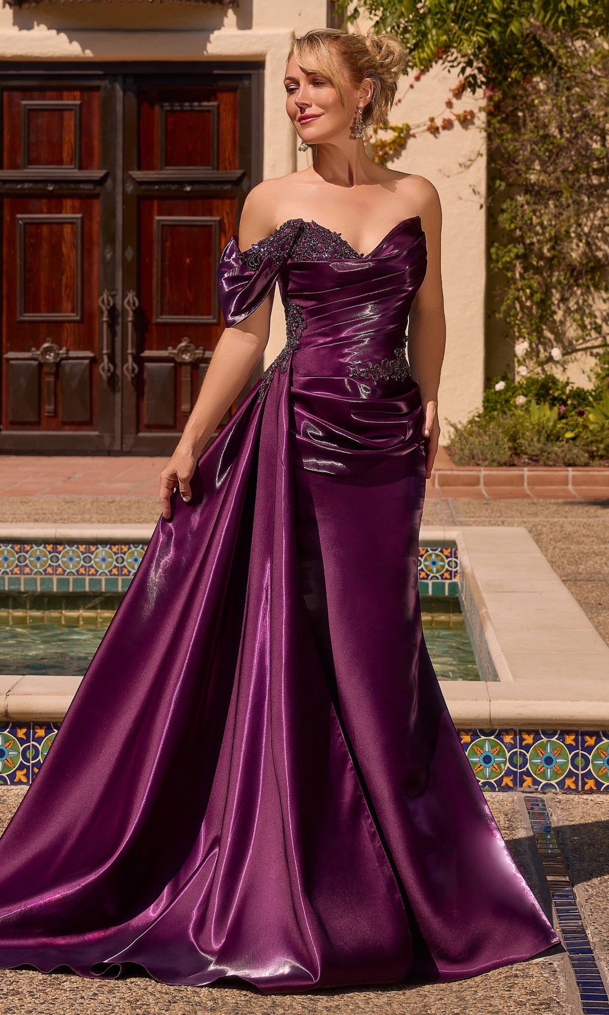 A woman wearing the Ladivine J916 long prom dress in a shiny eggplant hue with an off-the-shoulder neckline and dramatic side drape, standing outdoors near a tiled fountain and wooden doors.