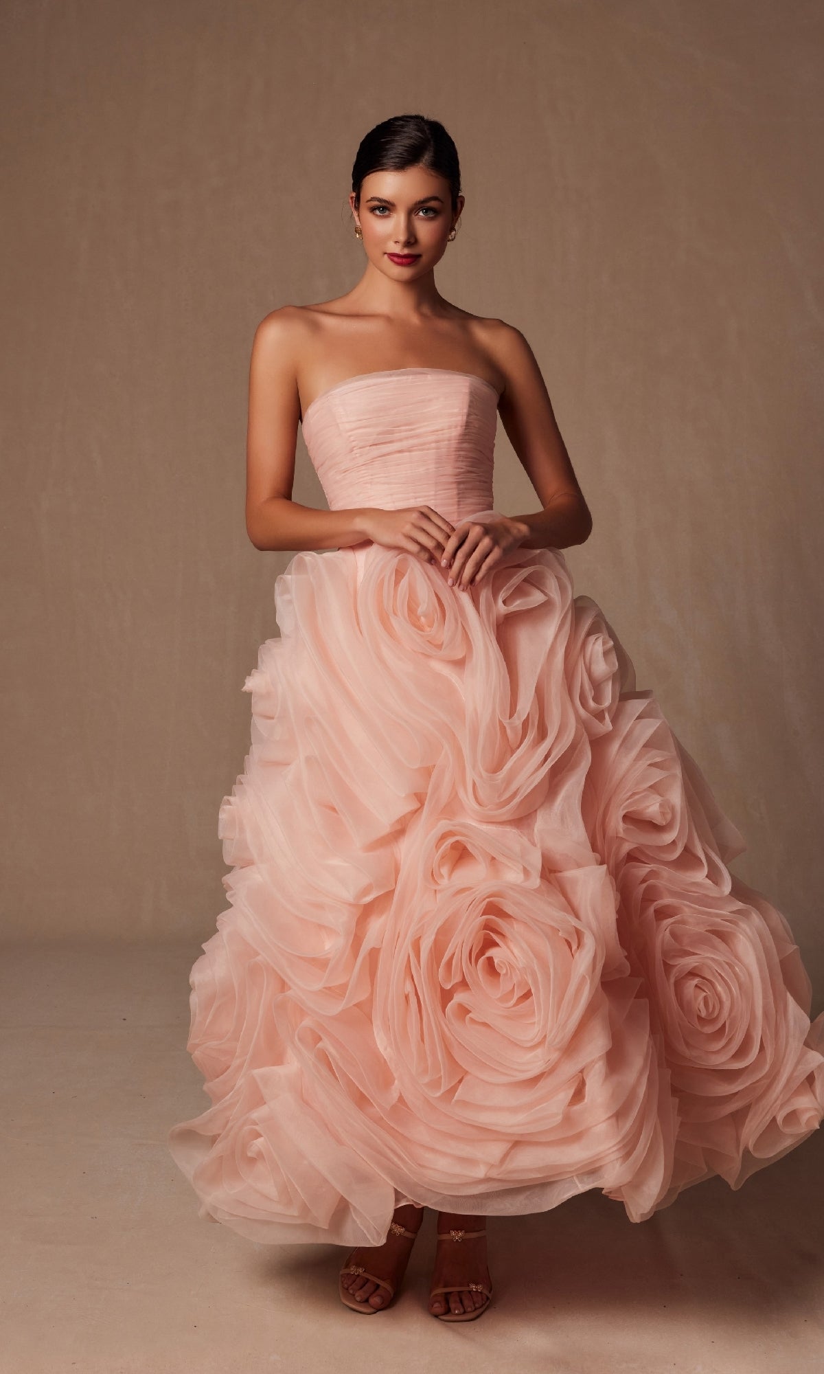 Woman wears the Nox Anabel JQ1037 strapless peach prom ball gown, featuring oversized rose-like ruffle rosettes, against a plain beige background.