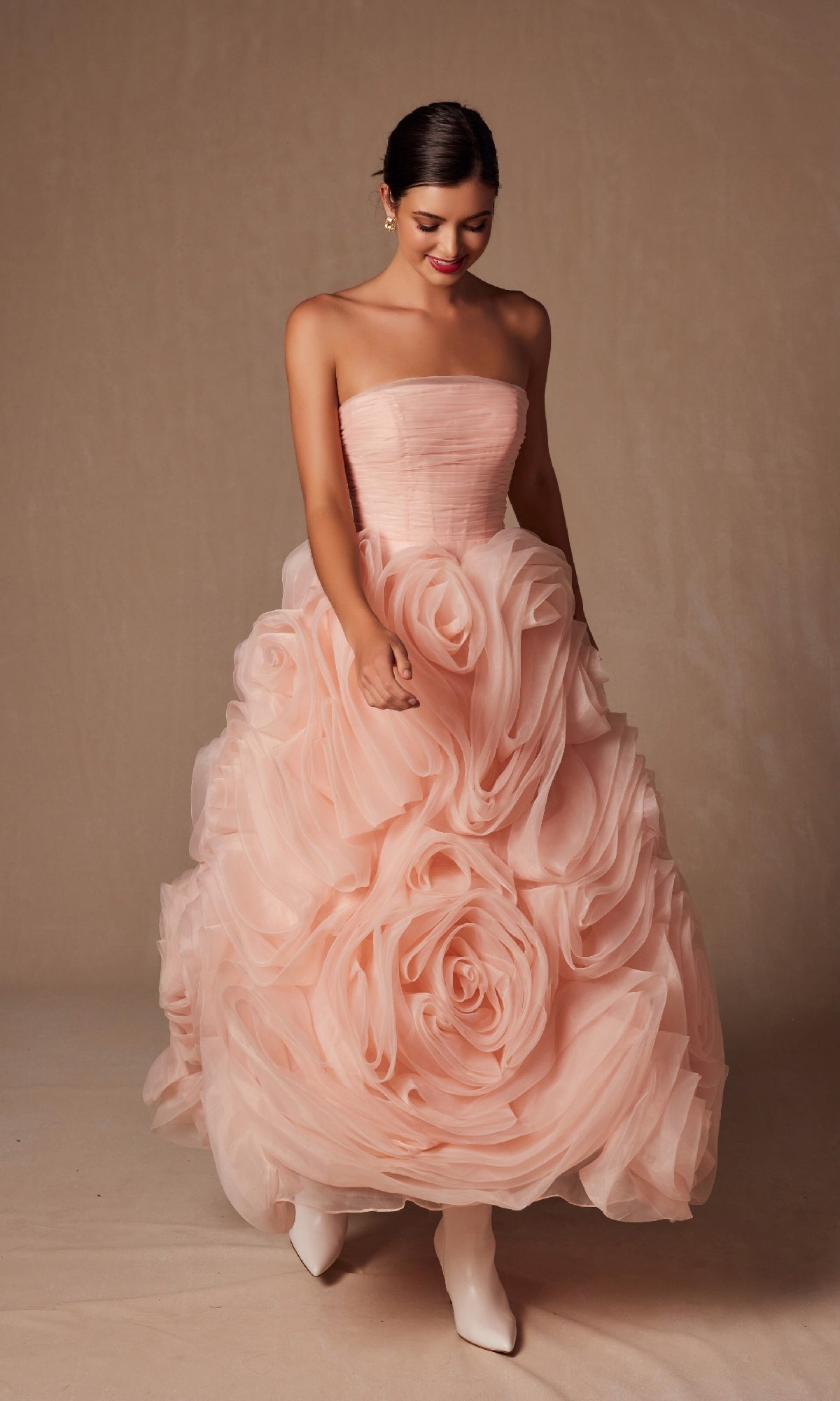 A woman in the Strapless Peach Prom Ball Gown (Nox Anabel JQ1037), featuring oversized rosettes on the skirt, stands against a beige background.