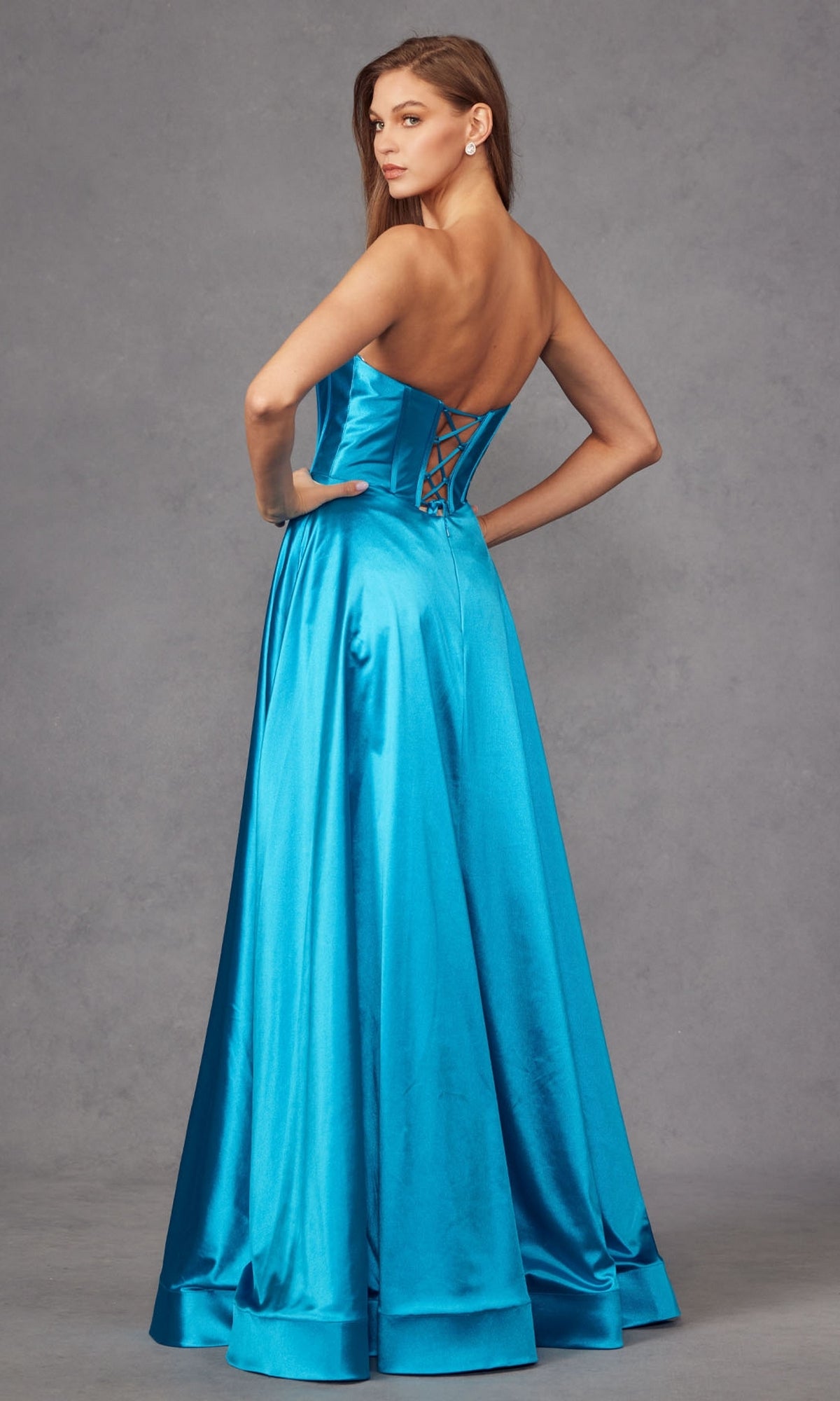 A woman models the Strapless Long Blue Prom Dress: Juliet JT2435A, featuring a sweetheart neckline, floor-length turquoise satin, and lace-up back, standing against a gray backdrop.