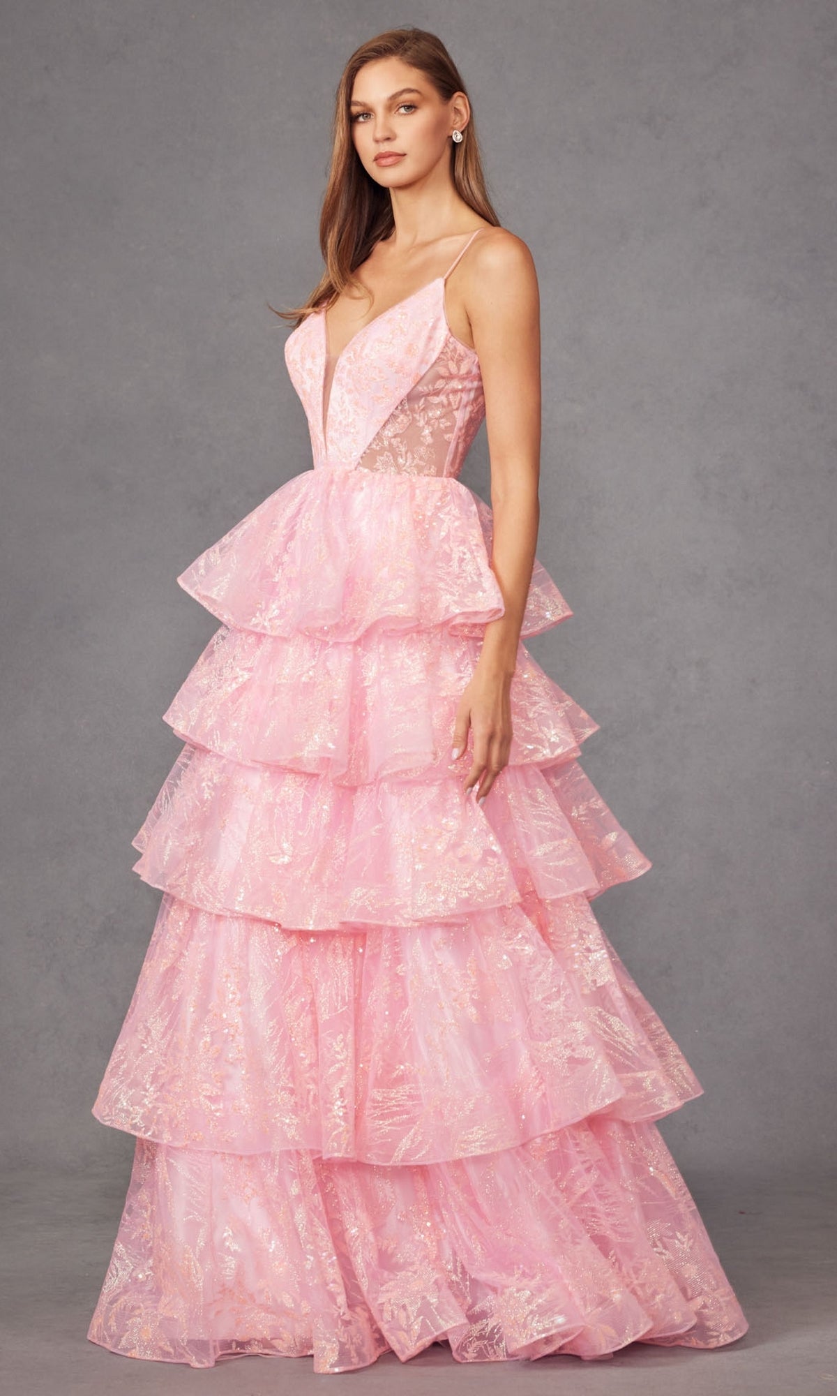 A woman poses in the Long Prom Dress JT2438K by Juliet, featuring a sleeveless deep V neckline, lace details, and tiered ruffle tulle layers, set against a plain gray background.