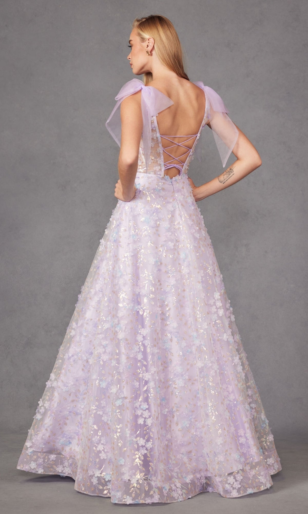 A woman stands facing away in the Juliet JT2439K, a lilac purple long lace prom dress featuring sequin floral details, sheer flutter sleeves, and an open back with lace-up detailing.