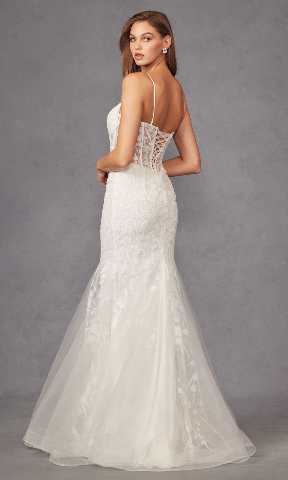 A woman stands before a gray background wearing the Off-White Long Prom Dress: Juliet JT2441SW, a mermaid-style gown featuring thin straps and a sheer corset back.