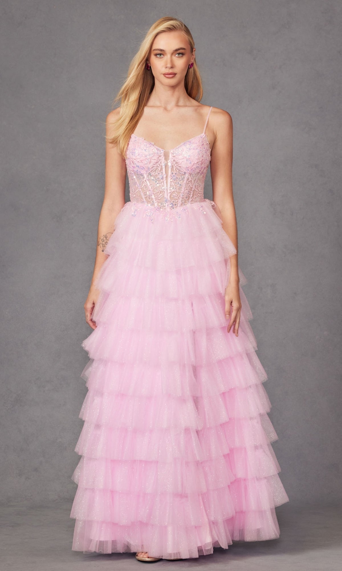 A woman stands against a plain gray background wearing the Juliet JT2458A Ruffled Glitter Tulle Prom Dress, featuring light pink tiers, spaghetti straps, and a semi-sheer embellished bodice.