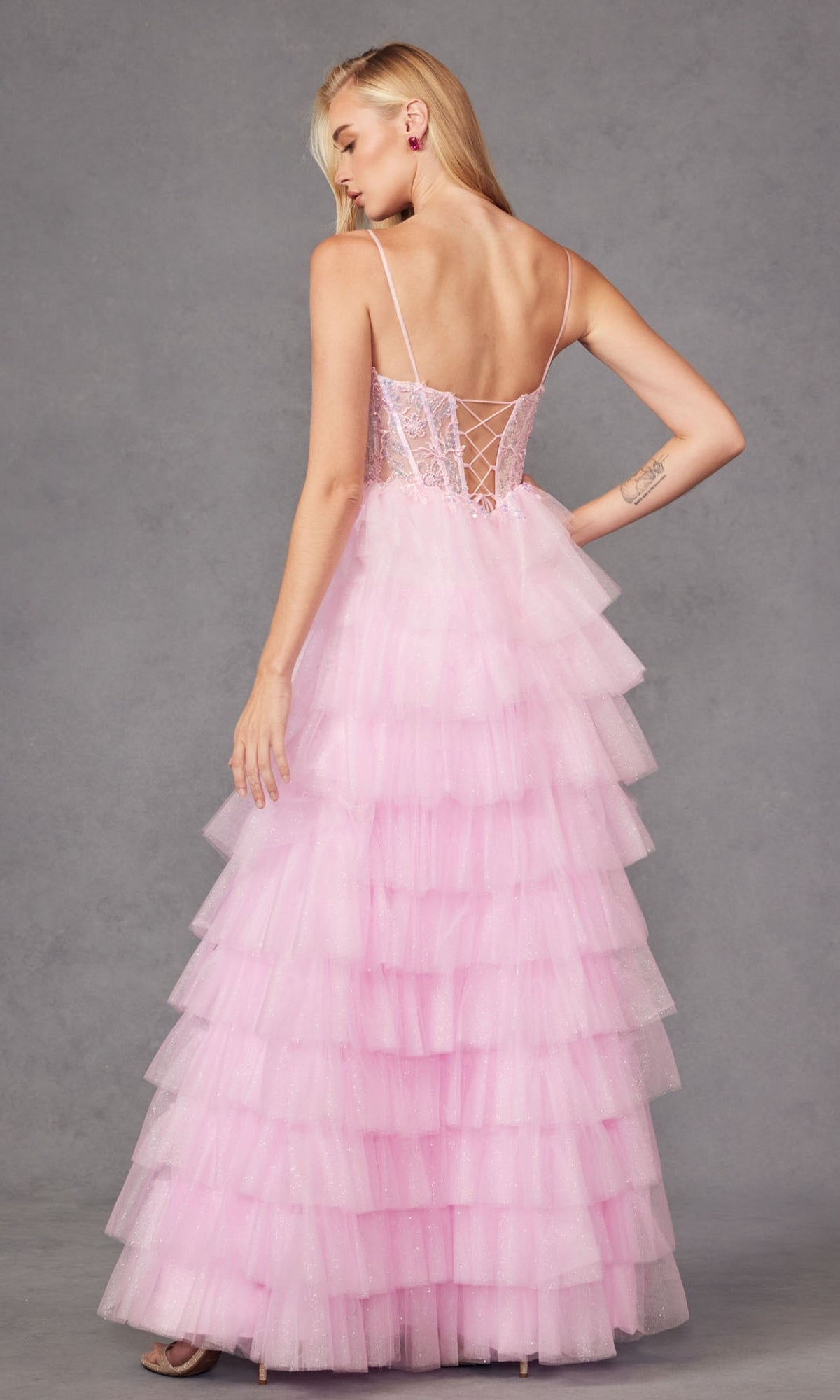 A person wears the Ruffled Glitter Tulle Prom Dress: Juliet JT2458A—floor-length, light pink with thin straps and a lace-up corset back—standing against a plain gray background.