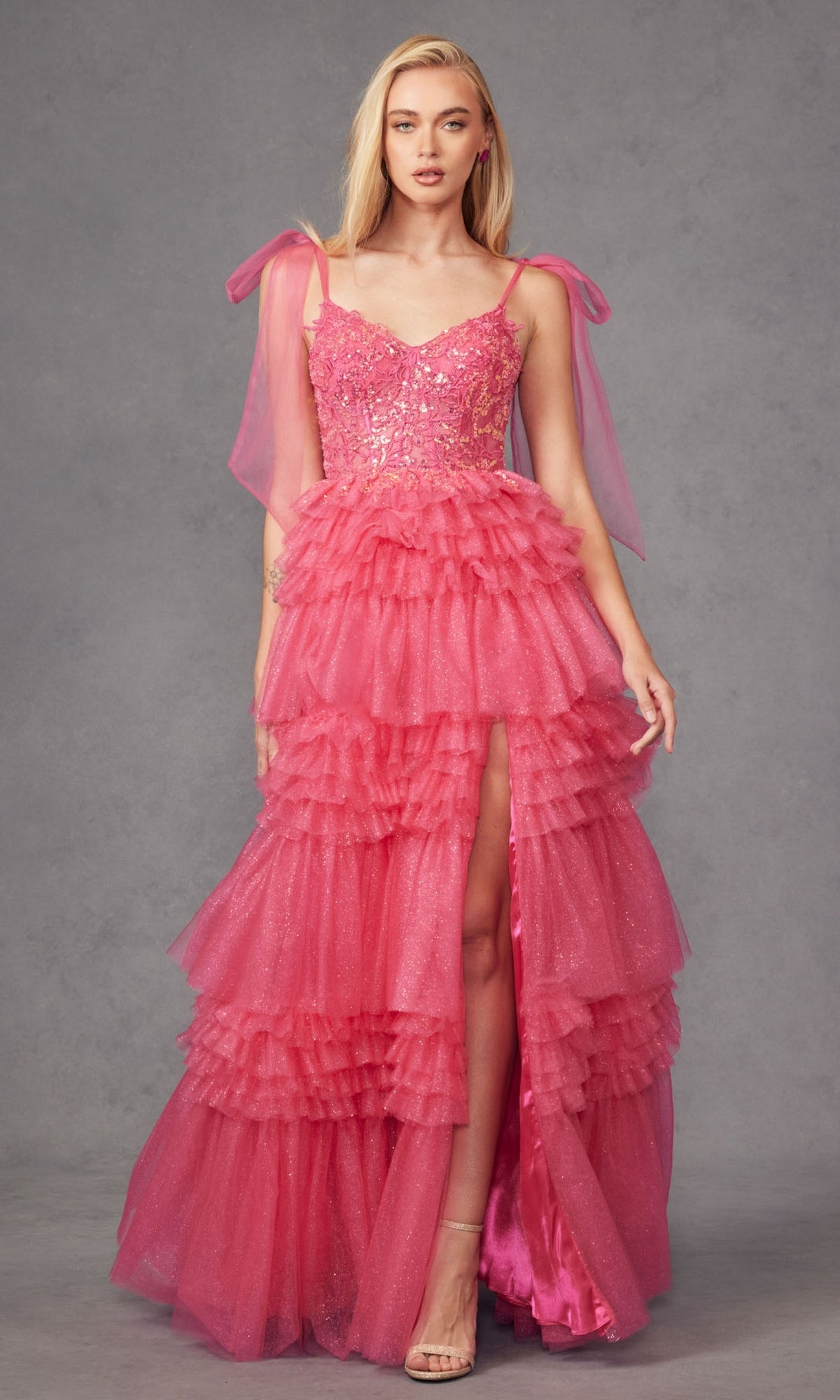 A woman in the Long Ruffled Prom Ball Gown: Juliet JT2465A, featuring bright pink tiered tulle, sequin embellishments, spaghetti straps with bows, and a thigh-high slit, stands against a gray background.