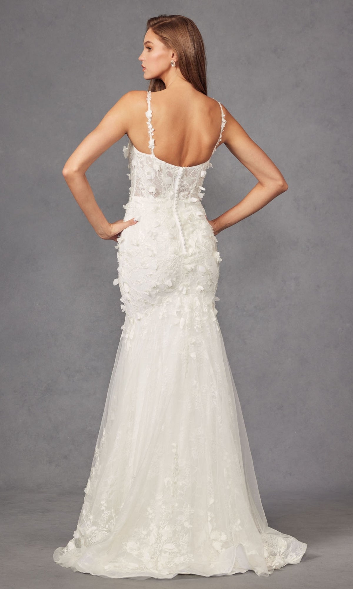 A woman stands facing away in the Long Prom Dress JT2475UW by Juliet, featuring an off-white mermaid silhouette with floral appliqués, thin straps, a low back, and an elegant train.