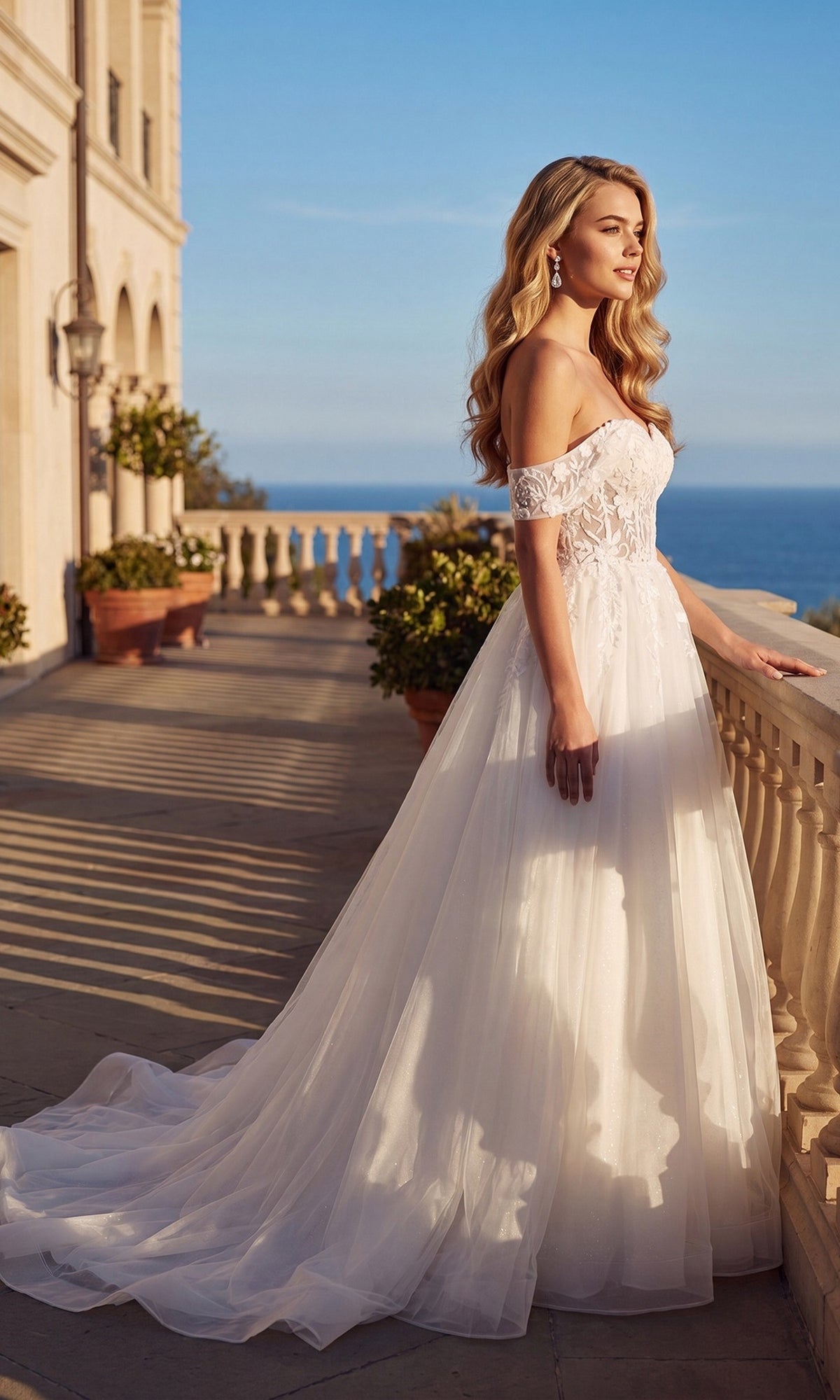 Long Off-White Bridal Dress: Juliet 82606L featuring an off-shoulder neckline, illusion lace bodice, and full tulle skirt, standing on a stone terrace overlooking the ocean.