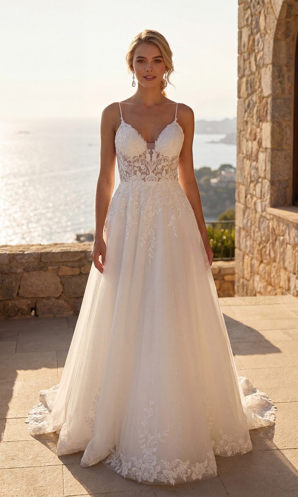 A woman stands outdoors on a stone terrace overlooking the sea, wearing the Juliet JT2633LW long formal dress in off white, a modern bridal gown with thin straps and elegant lace detailing.