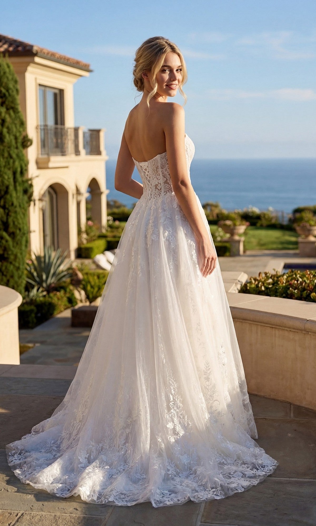 Wearing the Long Prom Dress: Juliet JT2670SW—a strapless lace formal gown with a sweetheart neckline—a woman stands outdoors on stone steps, glancing over her shoulder, with a garden, large house, and ocean in the background.