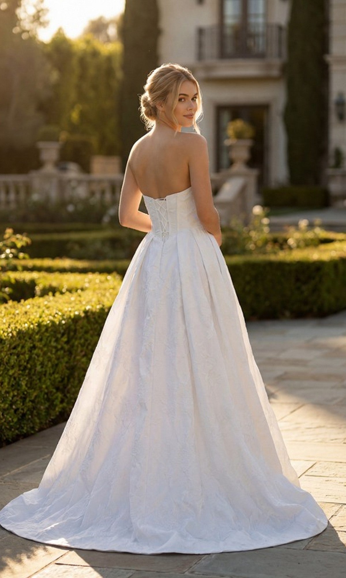 Wearing the Long Formal Dress: Juliet JT2692AW, a woman stands outdoors on a stone path, looking over her shoulder with a garden and large building in the background.