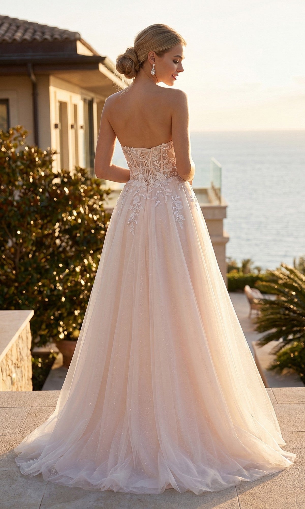 A woman wearing the Juliet JT2702AW long prom dress, a strapless floor-length blush gown with a delicate floral lace corset, stands outdoors facing the ocean, her hair styled in a low bun.