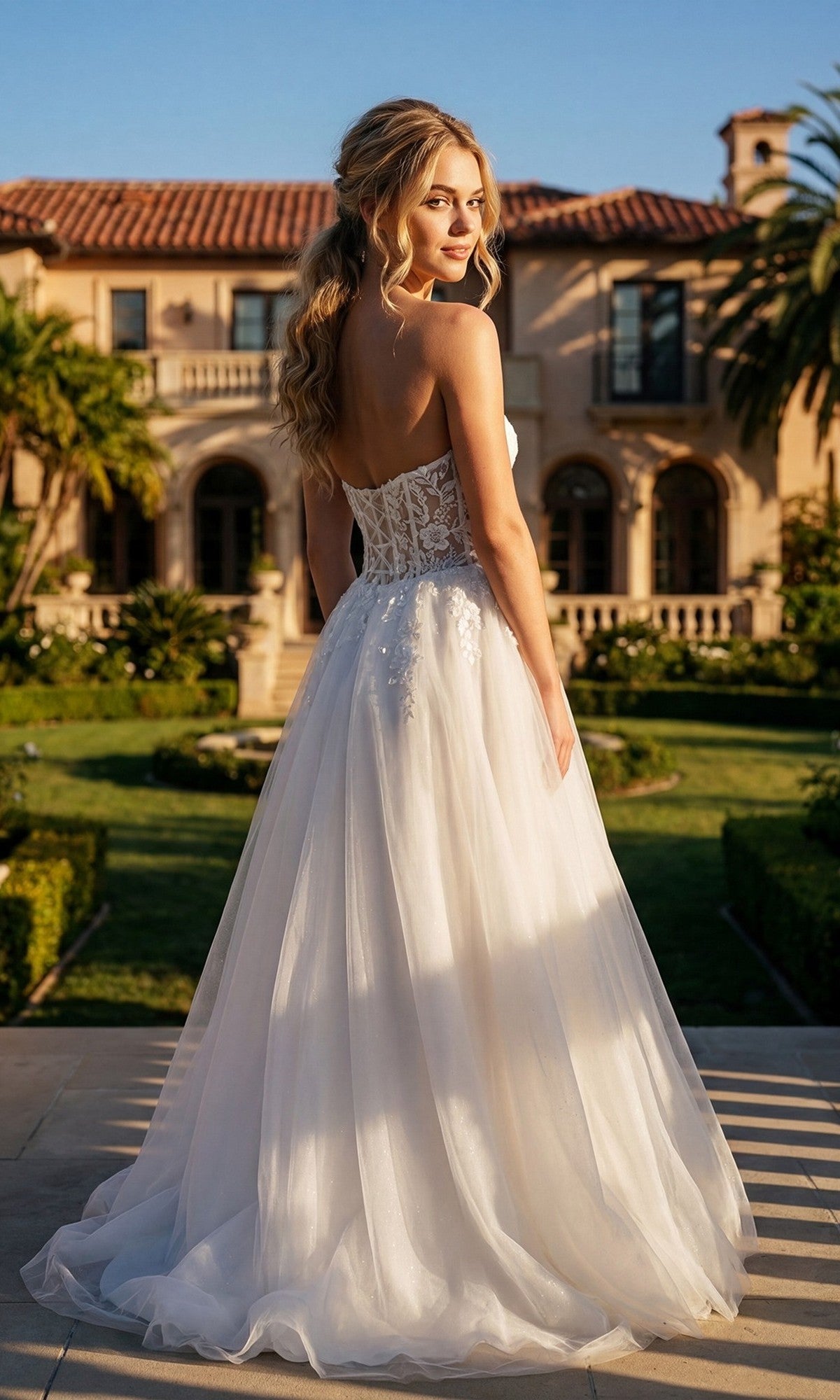 A woman in the Juliet JT2702AW long prom dress stands outdoors in front of a grand villa, looking over her shoulder as sunlight bathes the elegant a-line tulle gown. The strapless white design perfectly complements the picturesque setting.