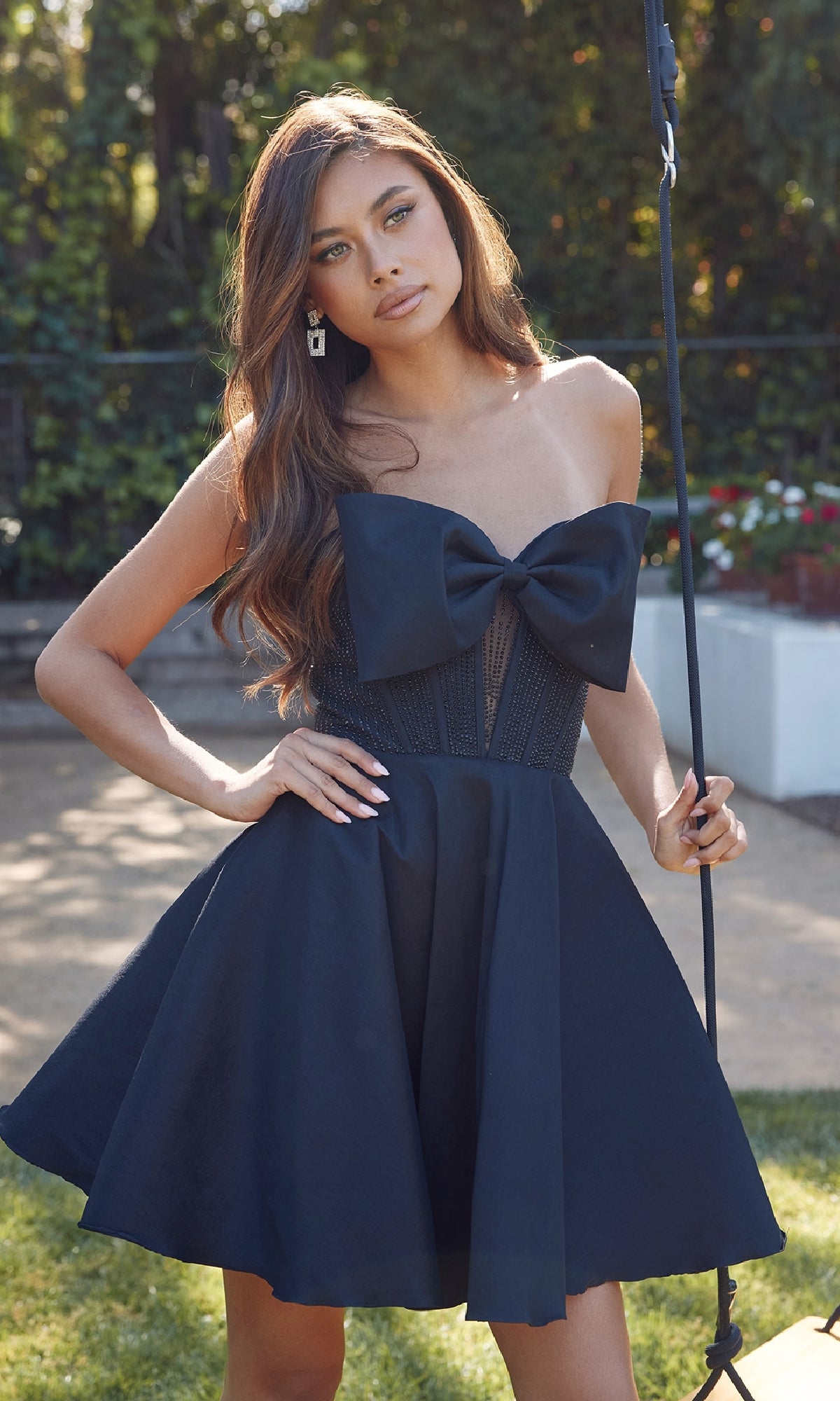 A woman stands outdoors in the Front Bow Strapless Hoco Dress: Juliet JT961A, featuring a large bow on the bodice and a flowing babydoll skirt, one hand on her hip and the other holding a black pole amid sunlit greenery.