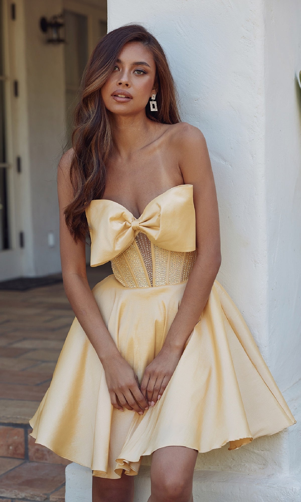 Woman in the Front Bow Strapless Hoco Dress: Juliet JT961A—a strapless, light yellow cocktail dress with a large front bow and flared babydoll skirt—stands outdoors against a white wall.