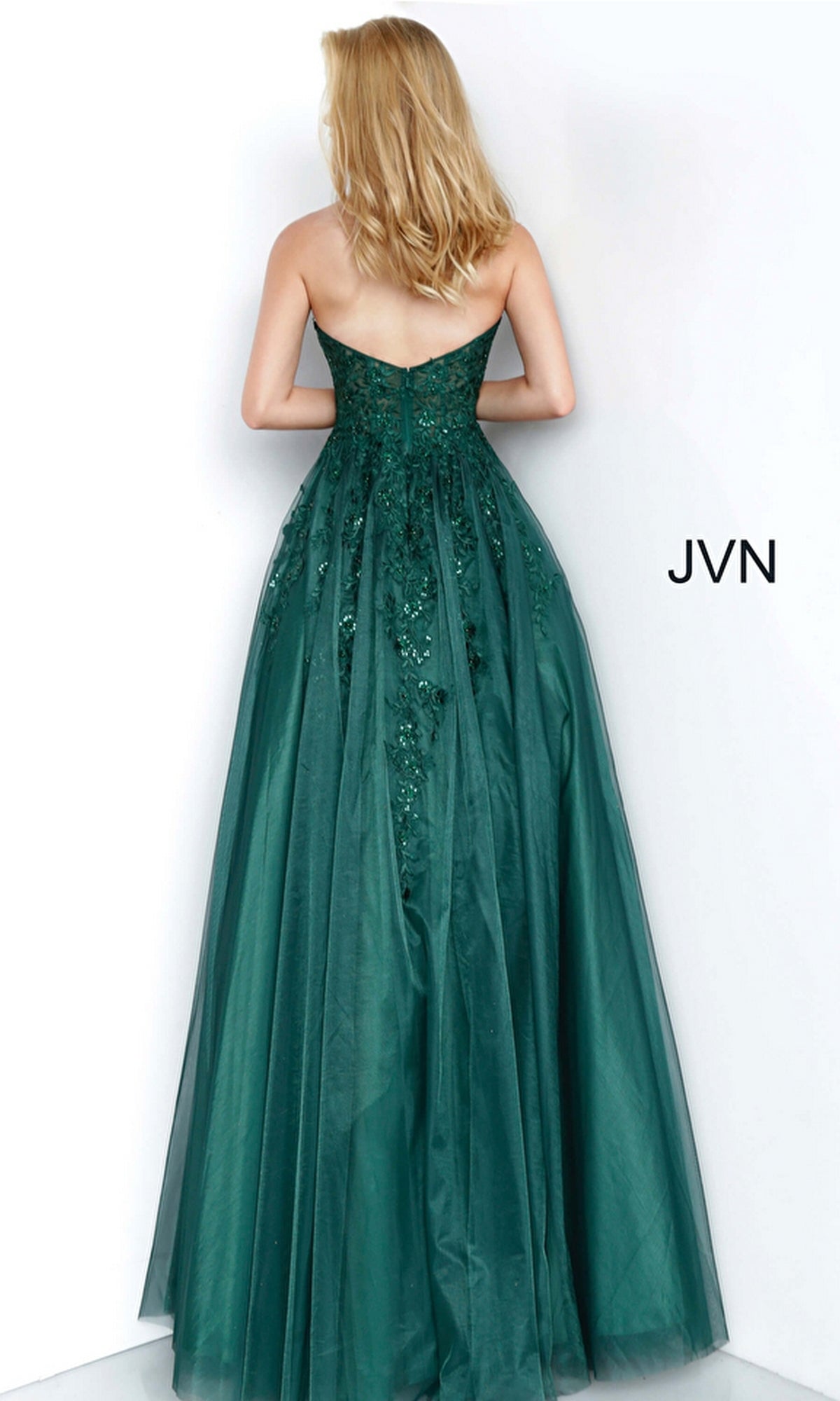 A woman stands facing a white wall wearing the Strapless Long Ball Gown: Jovani 00915—an emerald green, floor-length dress with floral embroidery and embellishments.