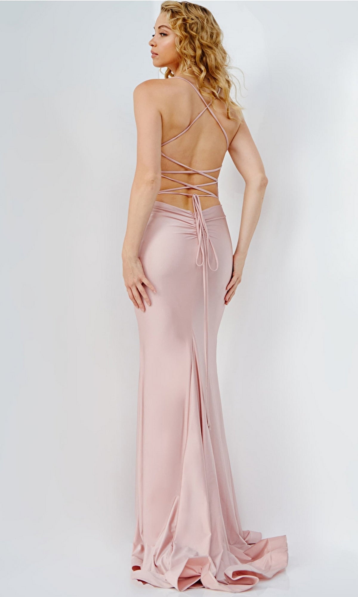 A woman stands with her back to the camera in the Lace-Up Jovani Long Prom Dress JVN09839, a light pink formal gown featuring crisscross straps, a backless design, and a slight train against a plain background.