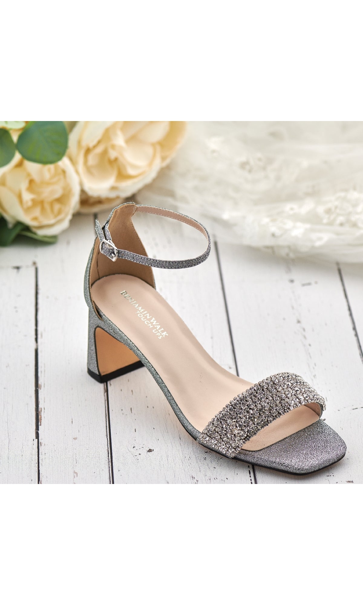 The Jade Pewter Prom Shoe (Touch Ups 4667) rests on a white wooden surface, featuring jeweled straps and an ankle buckle, surrounded by cream roses and delicate lace.