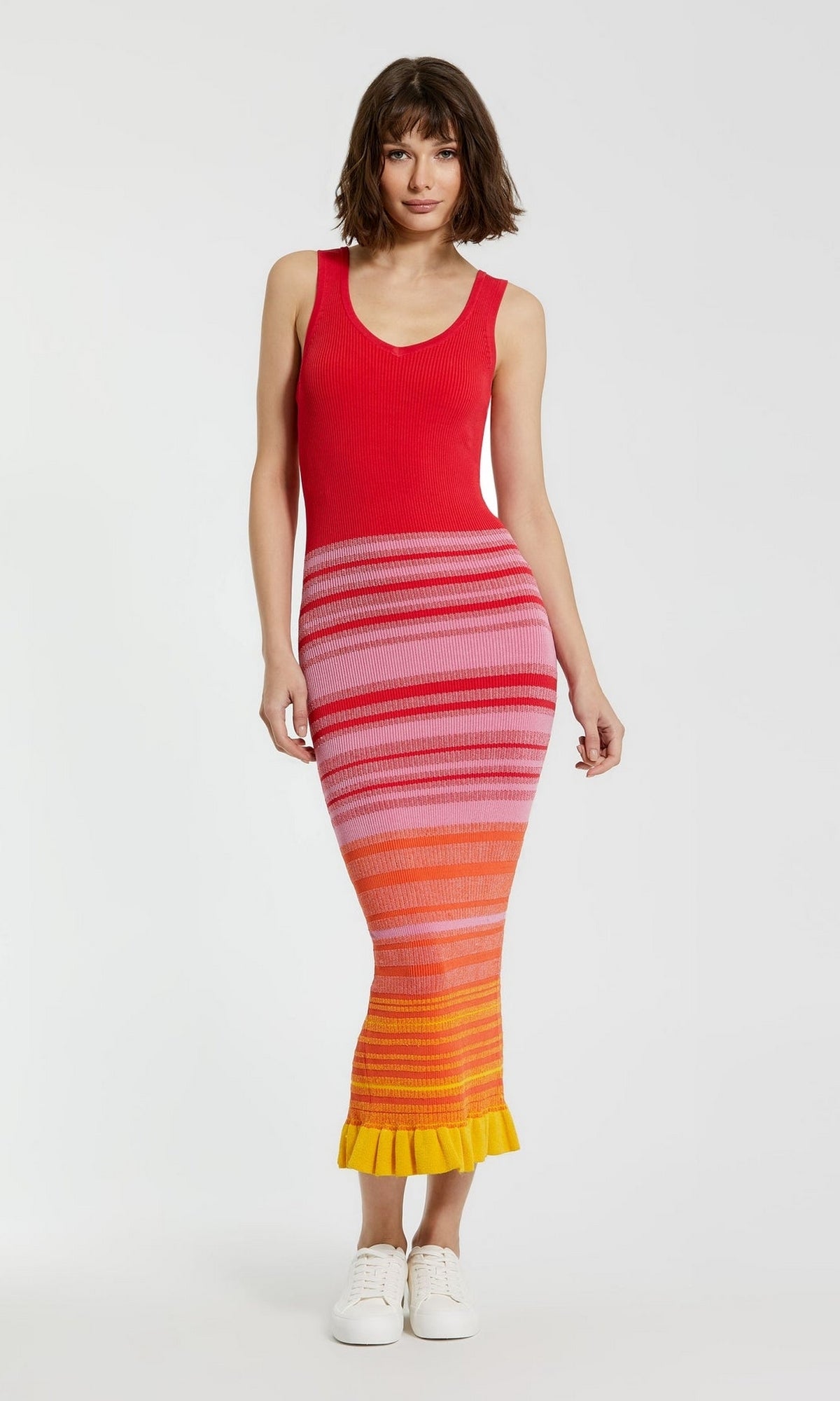 A woman stands against a plain white background wearing the Mac Duggal Red Casual Wedding-Guest Dress K12007, a striped tank dress in red, pink, orange, and yellow hues, paired with white sneakers.