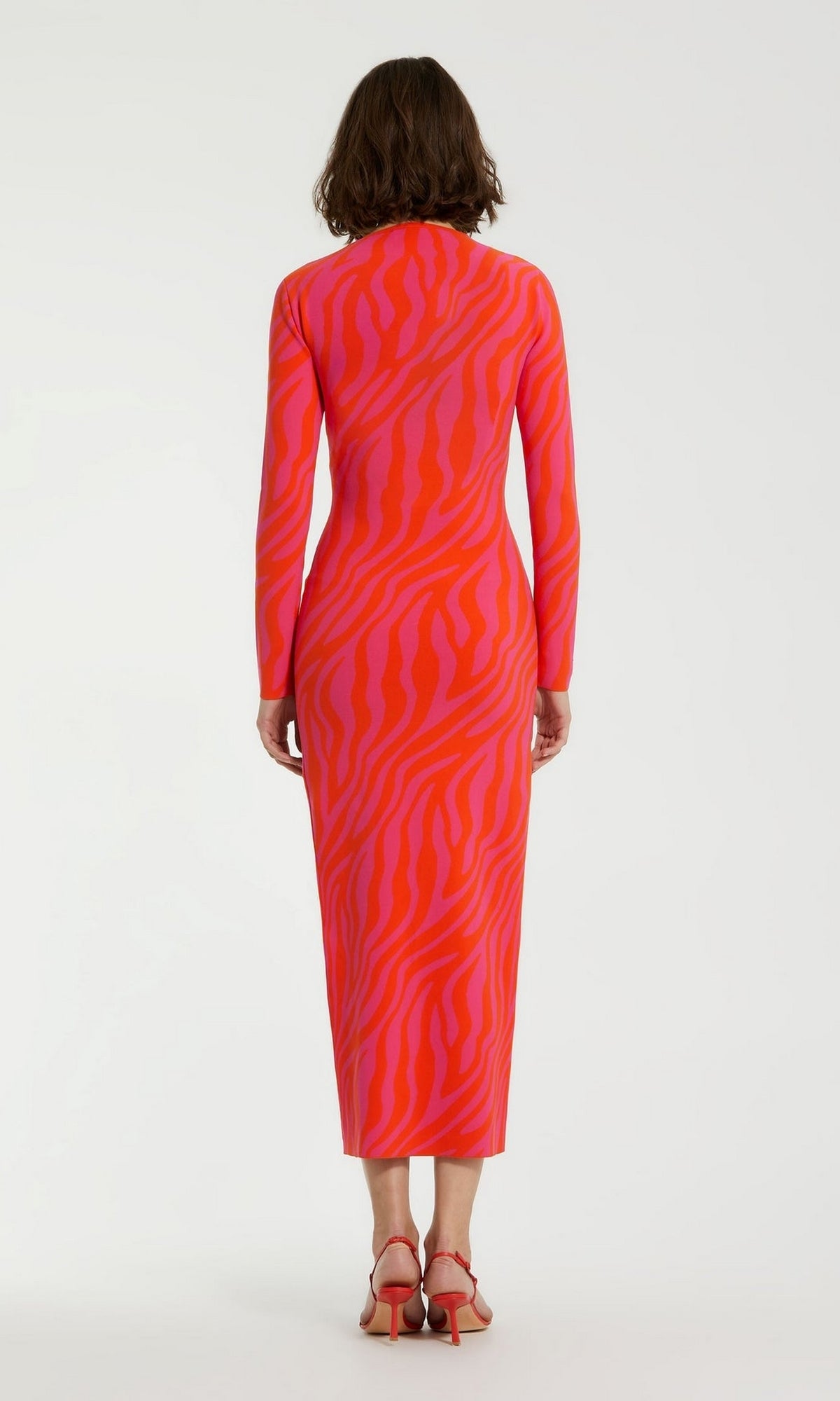 A person stands facing away, wearing the Mac Duggal Pink Casual Wedding-Guest Dress K12008, a long-sleeved, ankle-length zebra-print dress with red heeled sandals against a plain background.