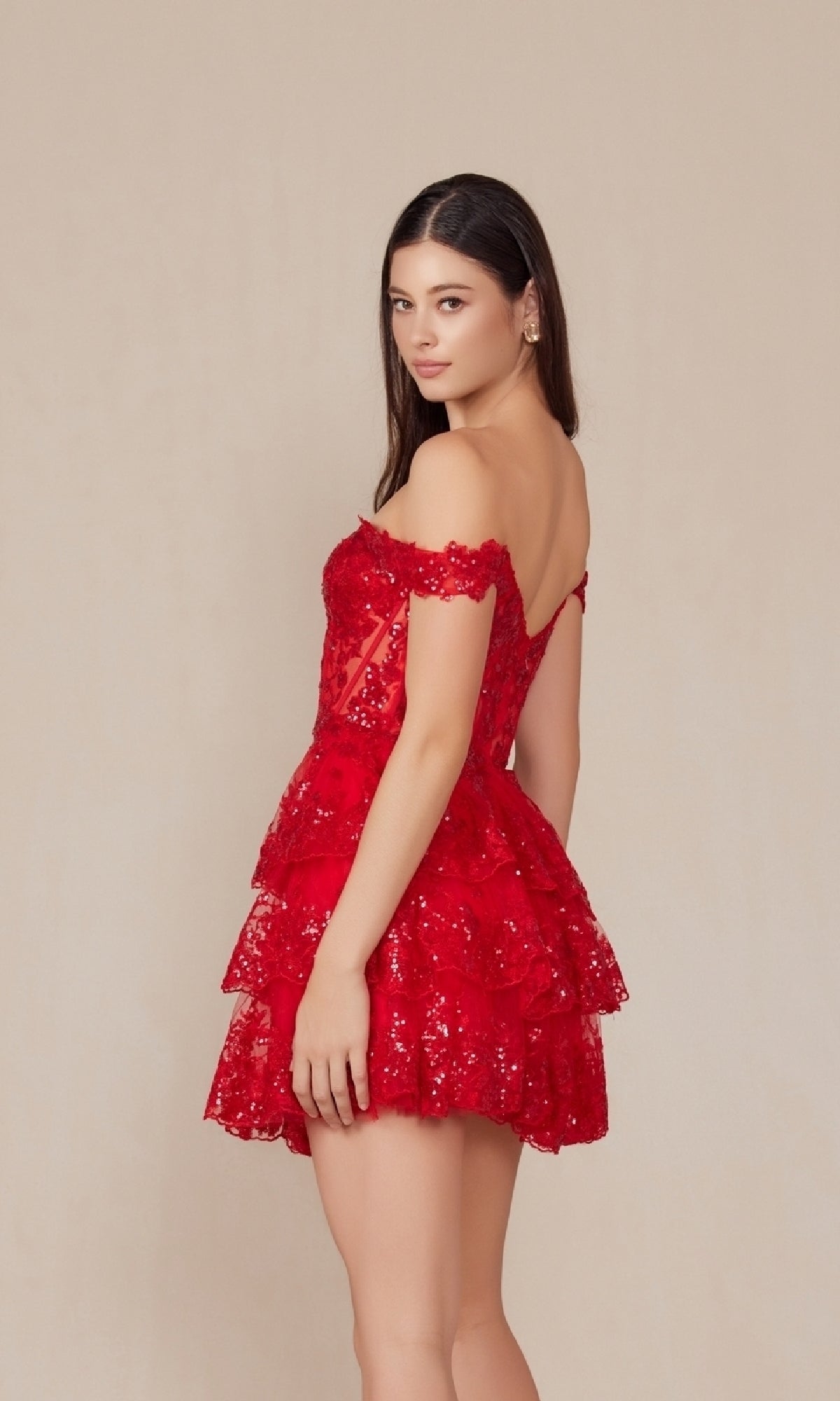 A woman stands sideways in the Off-Shoulder Short Ruffled Sequin Hoco Dress K856—a tiered, short red lace party dress with sequin details—set against a plain beige background.