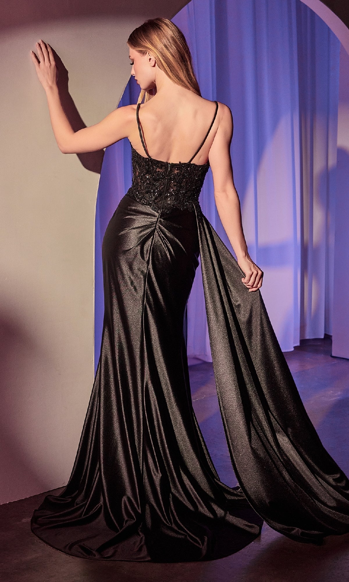 Wearing the Ladivine KV1117 Side-Sash Long Satin Prom Dress, a woman stands with her back to us, gracefully holding the train of her elegant gown, which features a beaded-lace bodice.