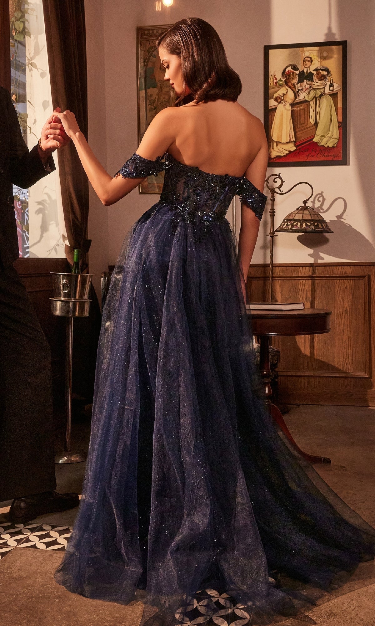 A woman indoors holds someone's hand, wearing the Ladivine KV1126 Beaded-Corset Long Prom Dress in dark blue, set against vintage decor and framed artwork.