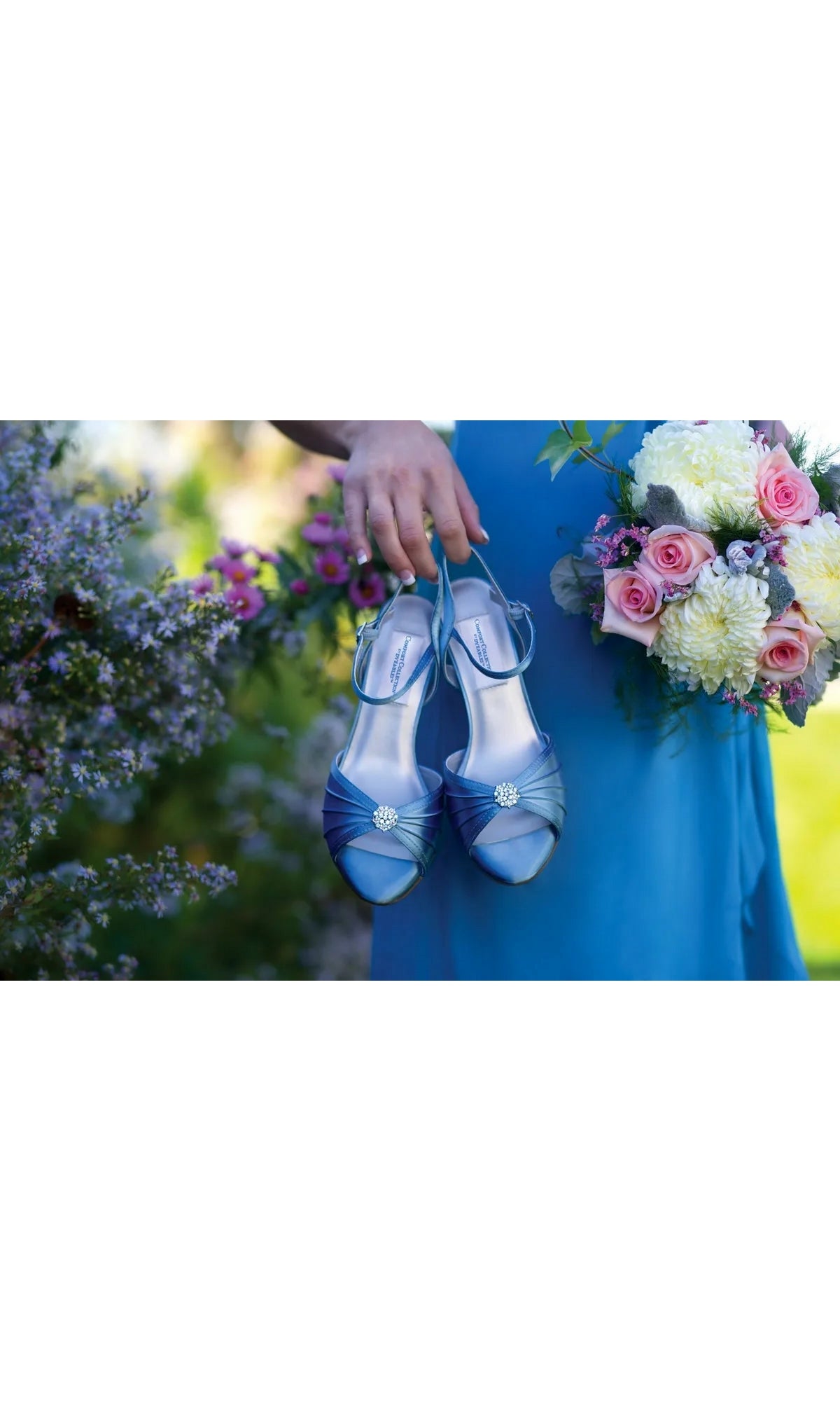 A person in a blue dress holds a pair of blue satin kitten heel peep-toe Kelsey White Prom Shoes: Dyeables 39114 with crystal accents next to a floral bouquet.
