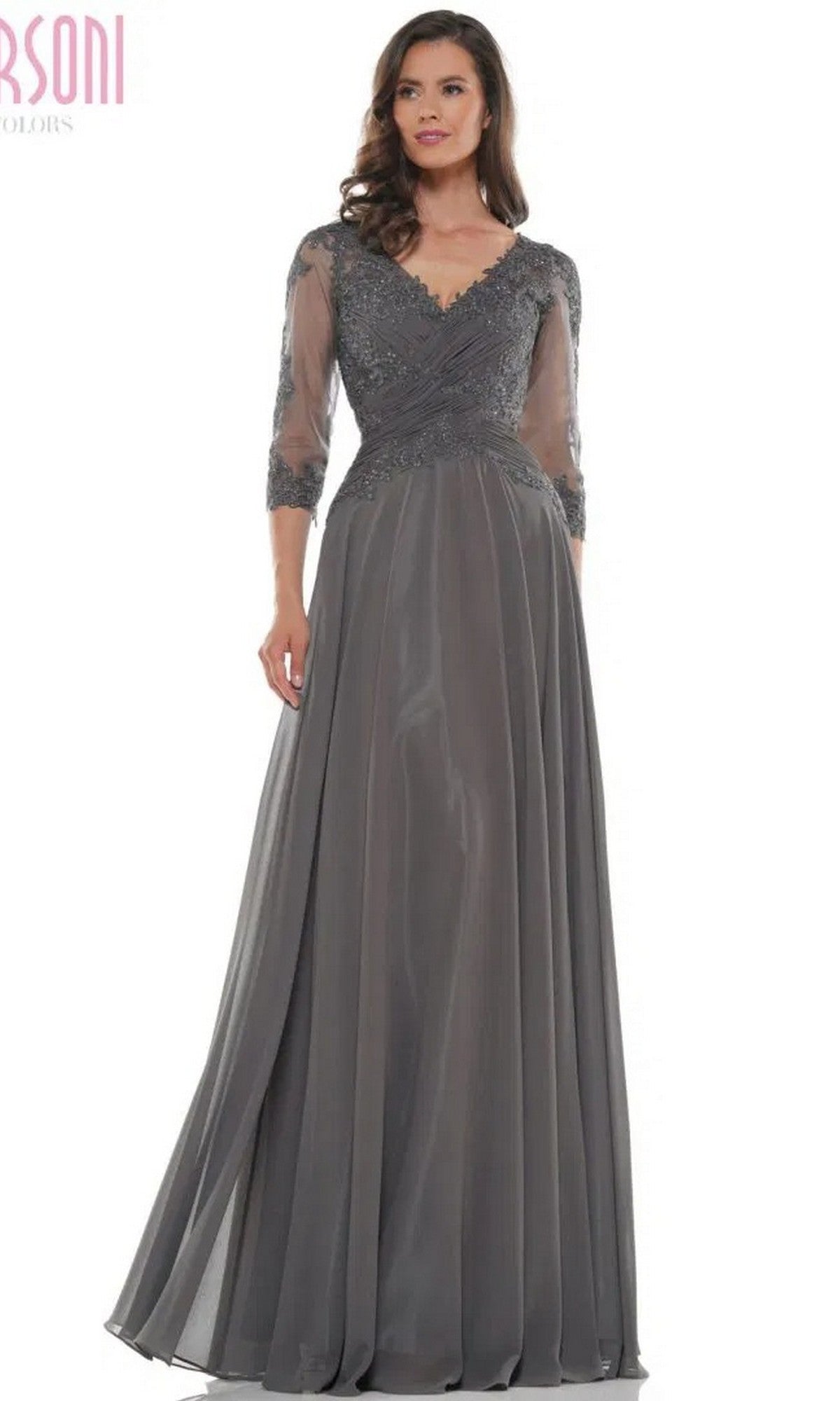 A woman in the Marsoni M237 long formal dress in charcoal grey, featuring an a-line silhouette, lace applique bodice, sheer three-quarter sleeves, and a V-neckline, stands against a plain background.