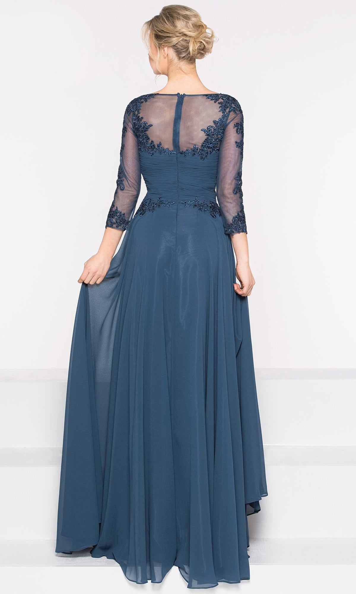 A woman in the Long Sleeve Long A-Line Formal Dress: Marsoni M237 faces away, holding the floor-length blue skirt with lace mesh sleeves and back.