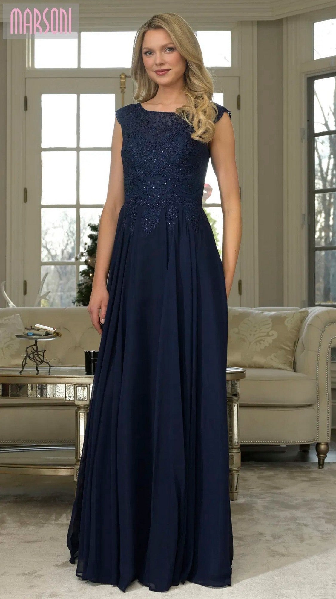 A woman stands indoors in front of a sofa and large windows, wearing the Marsoni M238 Long Formal Dress in navy with a fitted chiffon skirt and lace bodice.