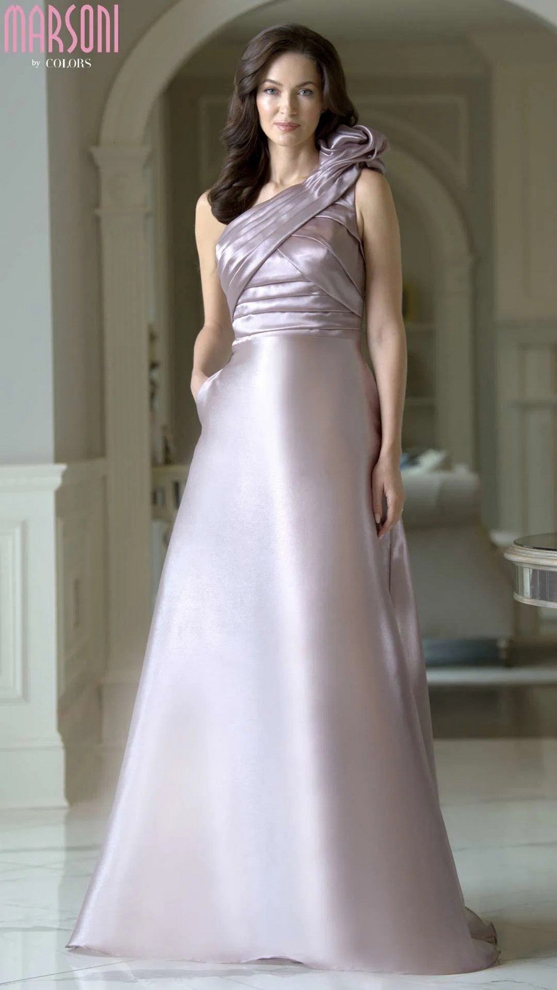 A woman stands indoors wearing the Marsoni M331 long formal dress in dusty rose—a shiny organza, one-shoulder gown with a floral strap detail and a graceful floor-length A-line skirt.