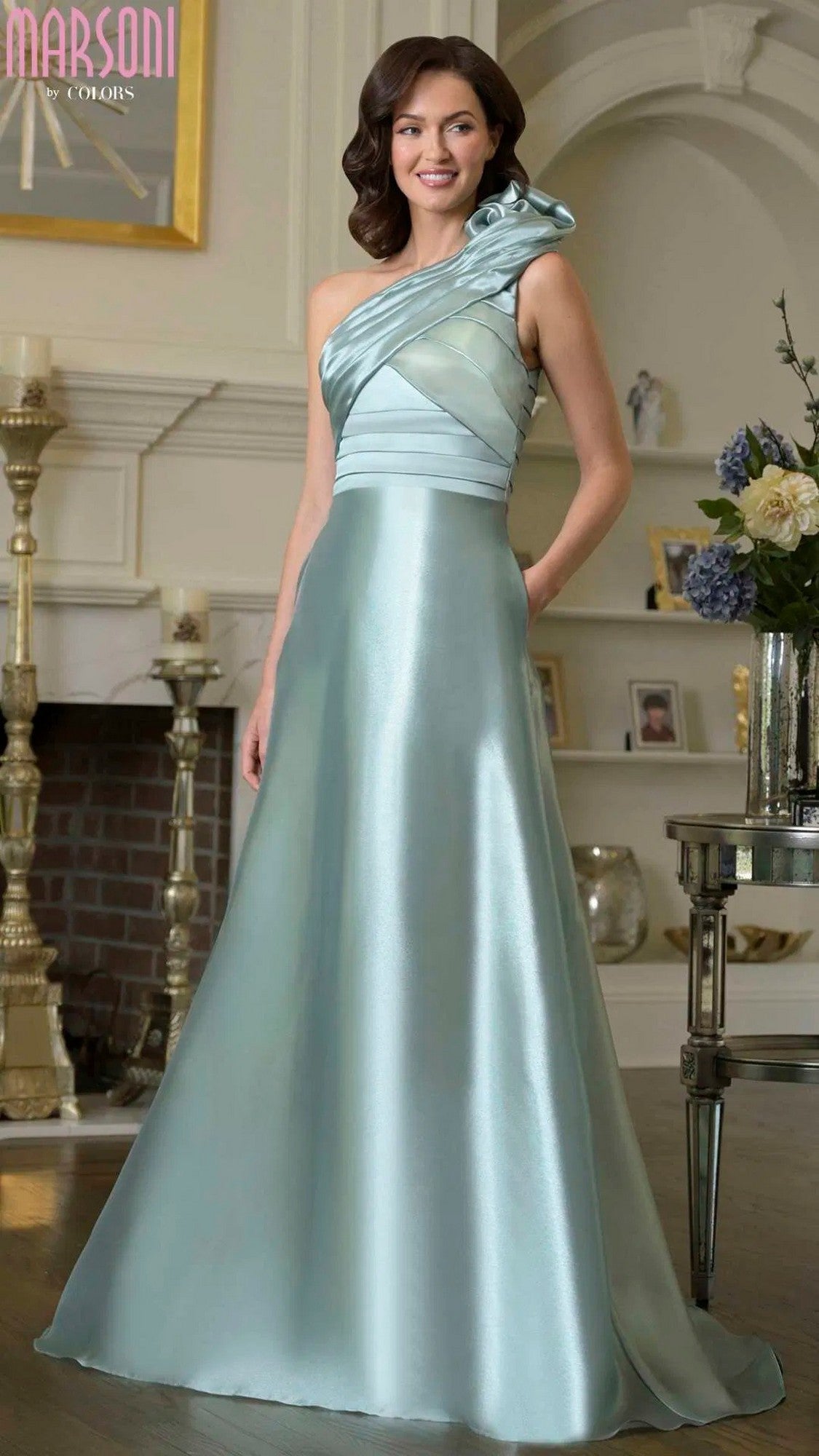 A woman stands indoors wearing the Marsoni M331, a sage one-shoulder long formal dress with a fitted bodice, shiny organza, and a flared, floor-length A-line skirt.