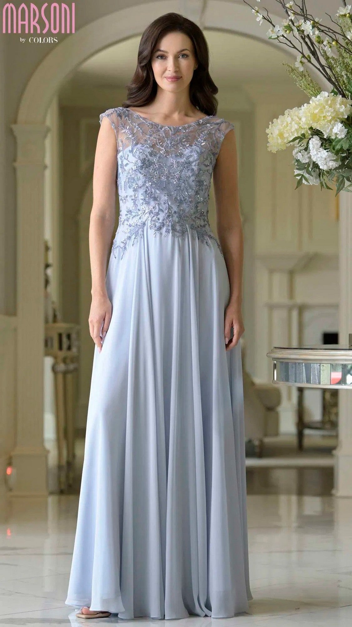 A woman wearing the Marsoni M340 Long Formal Dress in Dusty Blue, featuring a lace-embroidered bodice and chiffon A-line skirt, stands in a bright, elegant indoor setting.
