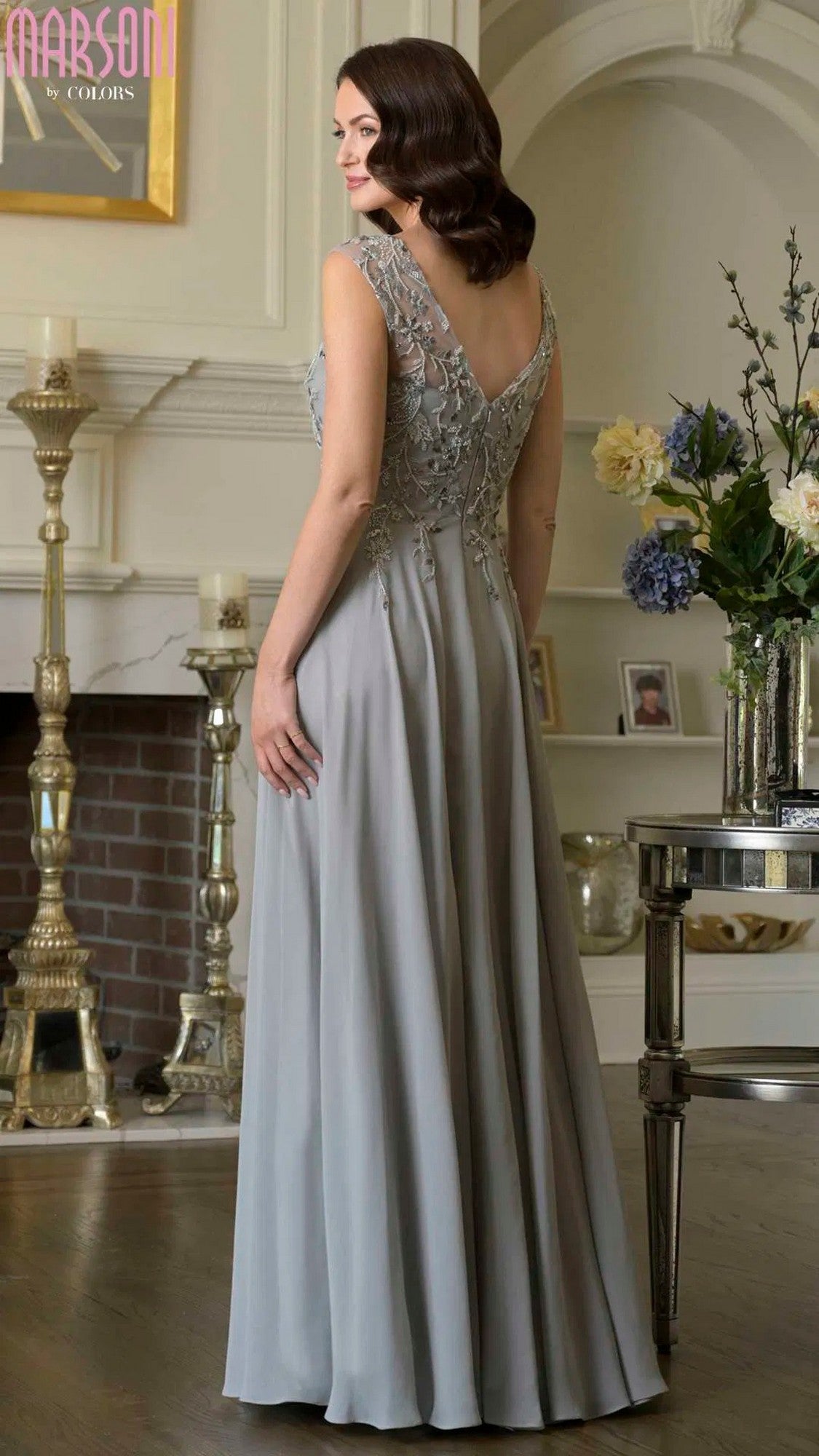 A woman wearing the Marsoni M340 long formal dress, featuring a silver-gray hue and lace-embroidered bodice, stands indoors near a fireplace and a table decorated with flowers and framed photos.