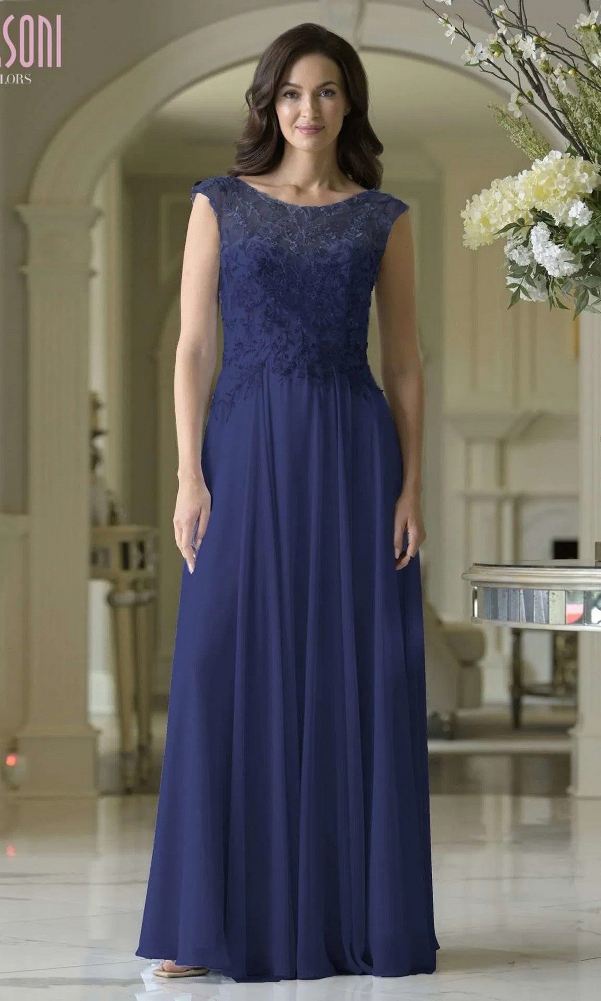 A woman stands indoors wearing the Marsoni M340 long formal dress in navy, featuring a sleeveless lace-embroidered bodice and an elegant chiffon A-line floor-length skirt.