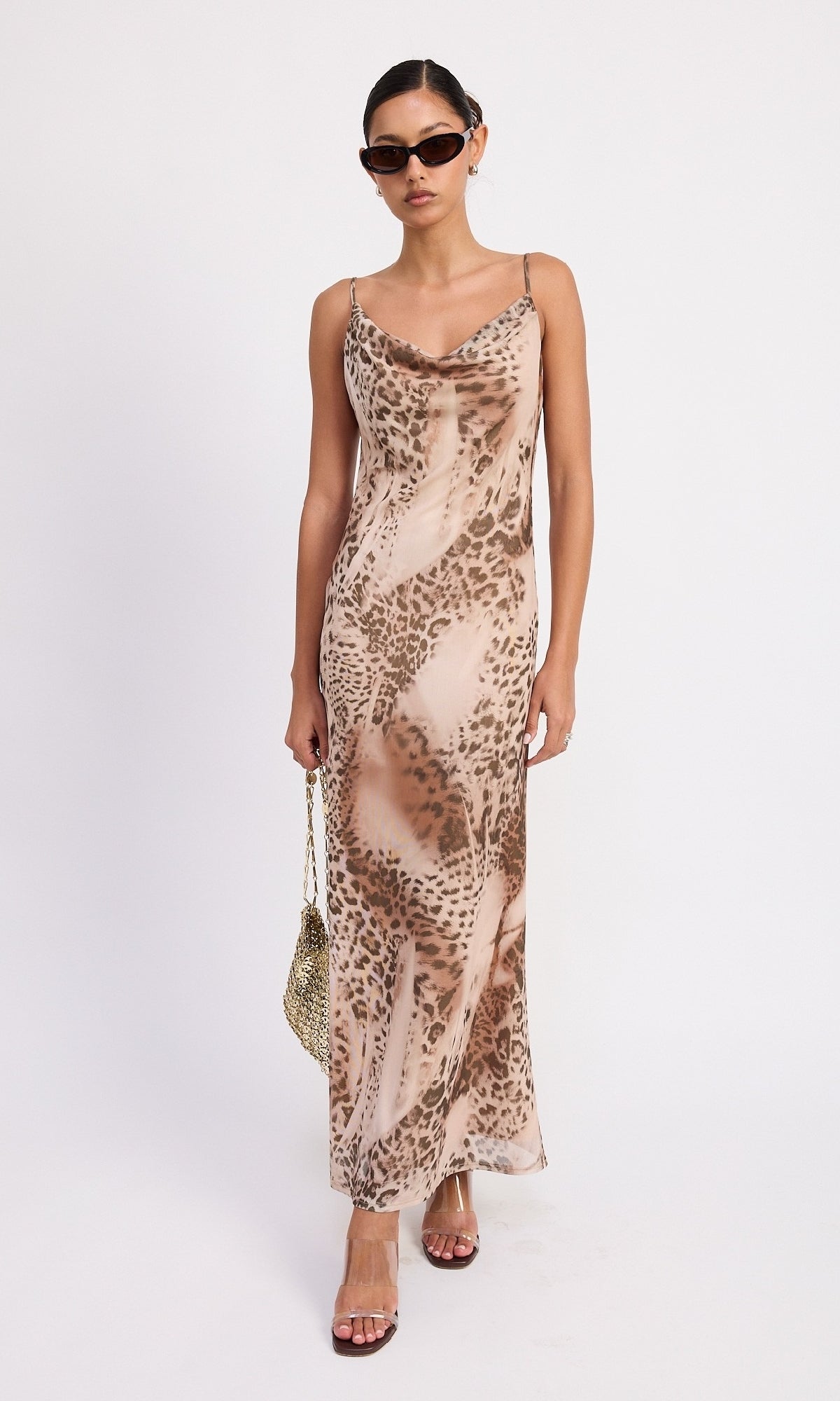 A woman wearing sunglasses and holding a small handbag models the Brown Leopard Cowl-Neck Slip Dress against a plain white background.