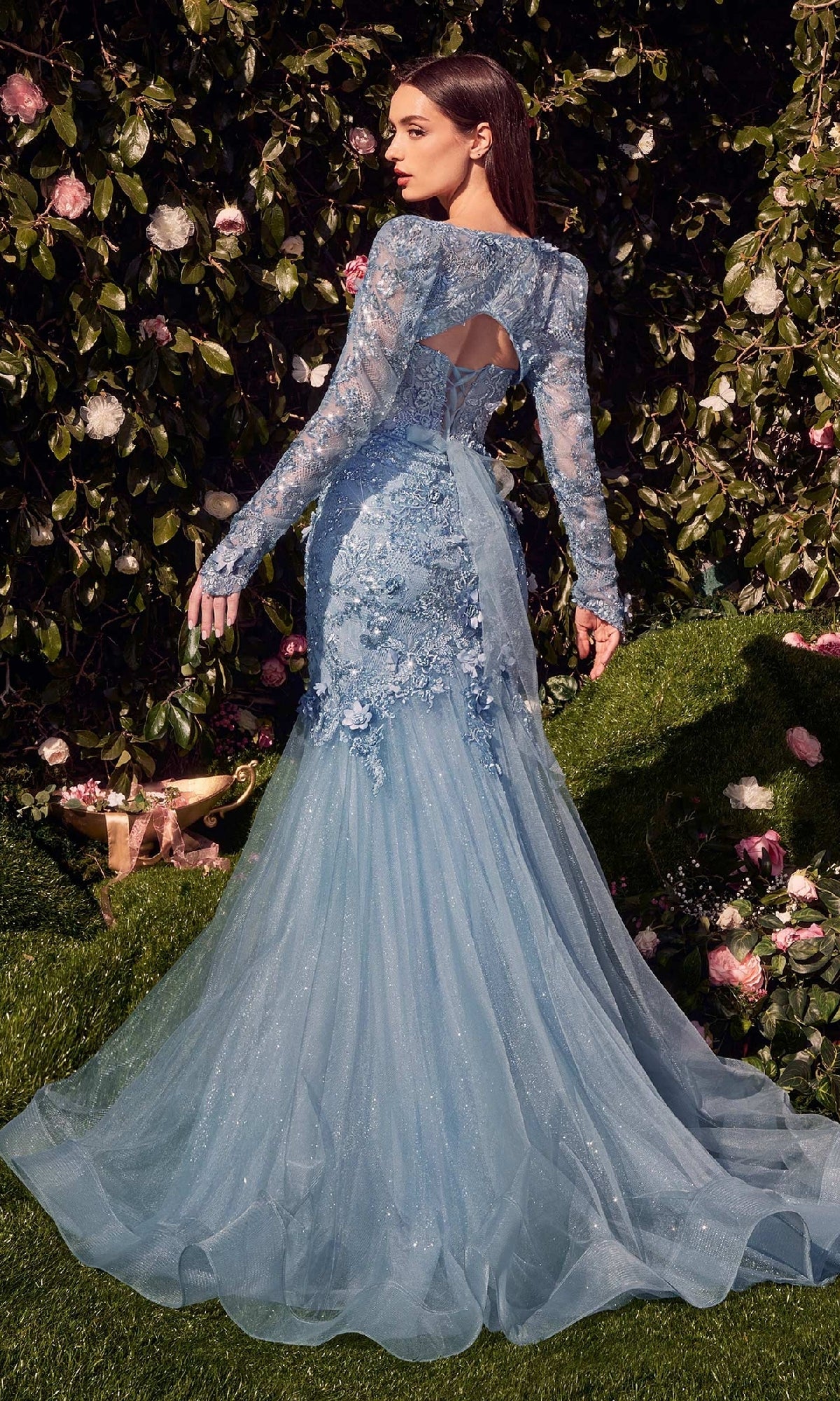 A woman in the Andrea & Leo MA101 long sleeve long formal dress with a beaded lace bodice and flowing tulle train stands outdoors by a bush with pink and white flowers.