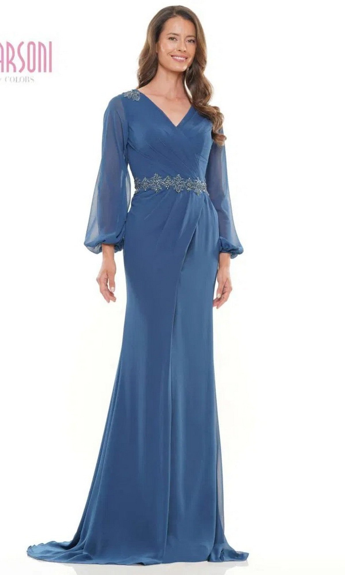 A woman smiles against a plain white background, modeling the Marsoni MV1273—an elegant, floor-length long sleeve formal dress featuring floral embroidery at the waist.