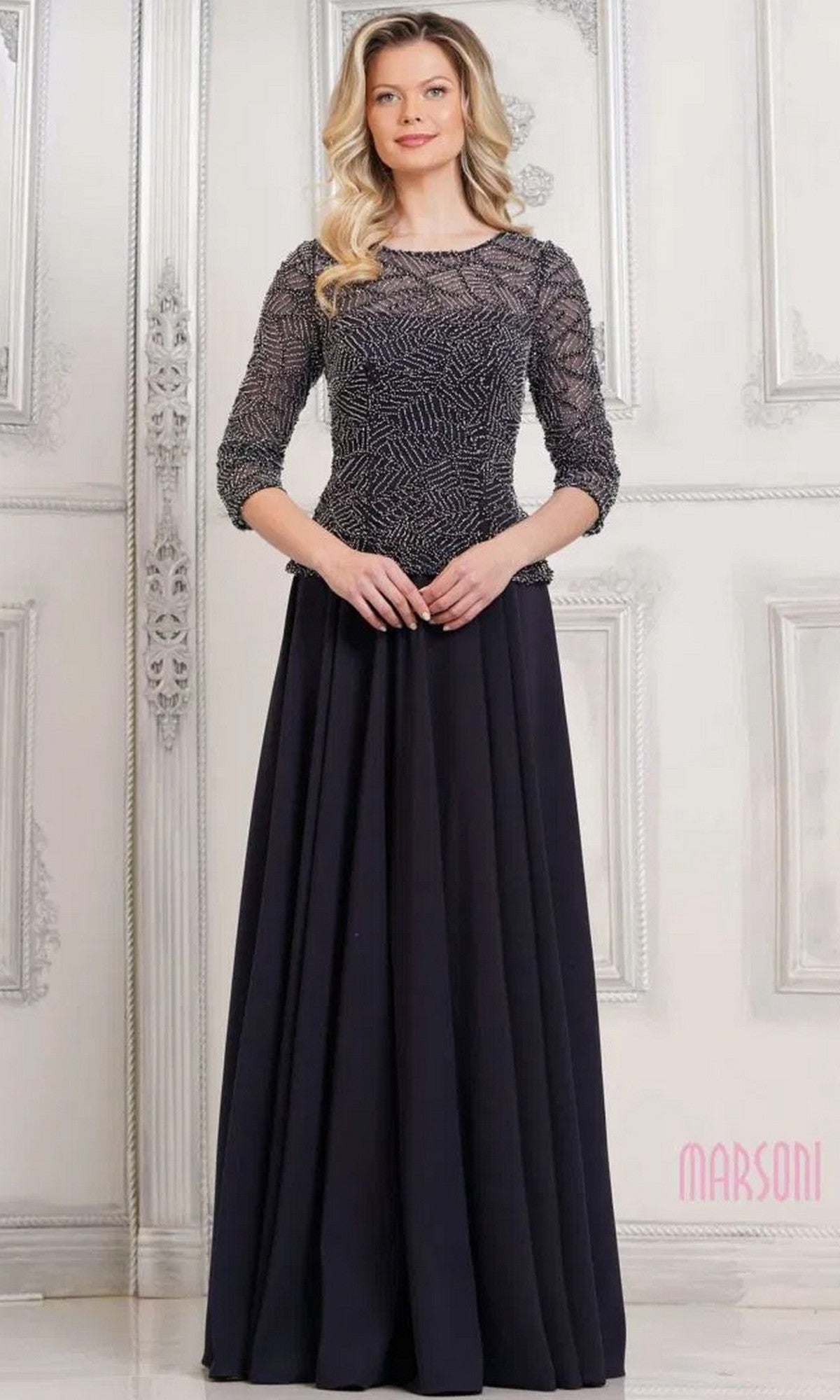 A woman poses in front of a white ornate wall wearing the Marsoni MV1319 Long Formal Dress in Midnight, featuring a beaded bodice, three-quarter sleeves, and a flowing A-line skirt.