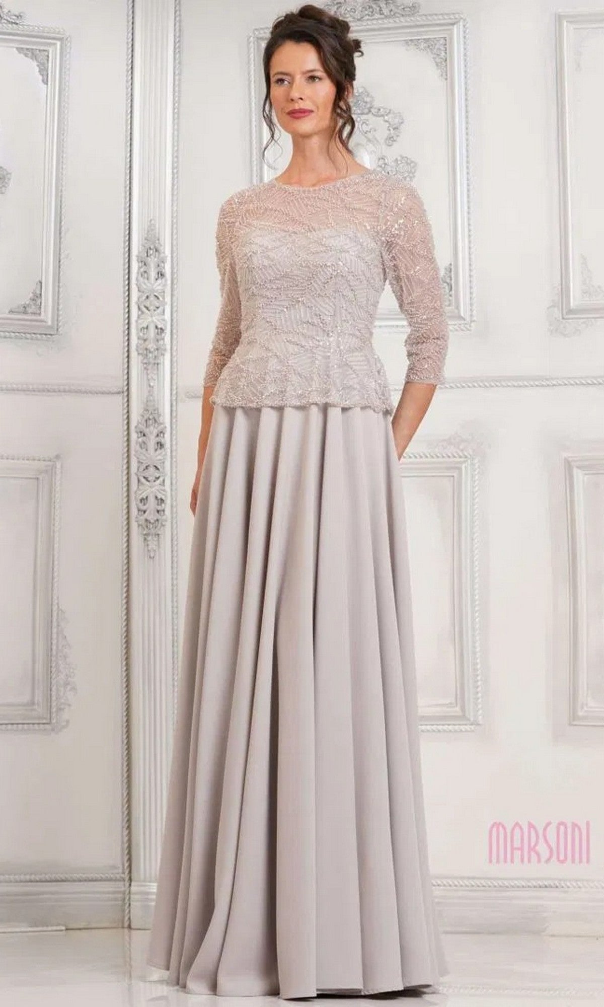 A woman wears the Marsoni MV1319 long formal dress in oatmeal, featuring a beaded bodice with three-quarter sleeves and an elegant A-line skirt, standing against an ornate white wall.