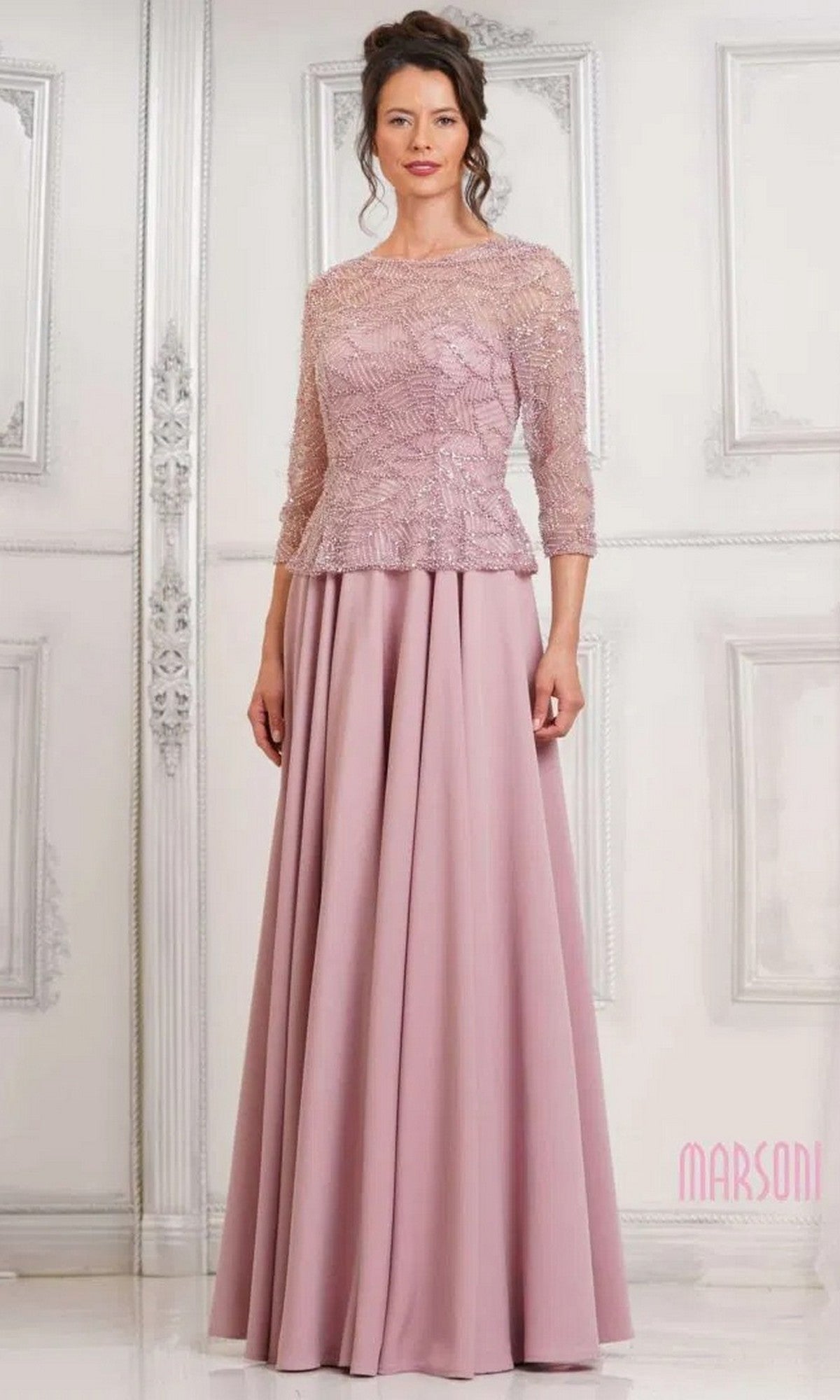 A woman wears the Marsoni MV1319 long formal dress in rose, featuring an elegant lace bodice, three-quarter sleeves, and a graceful A-line skirt, as she stands before a white paneled wall.