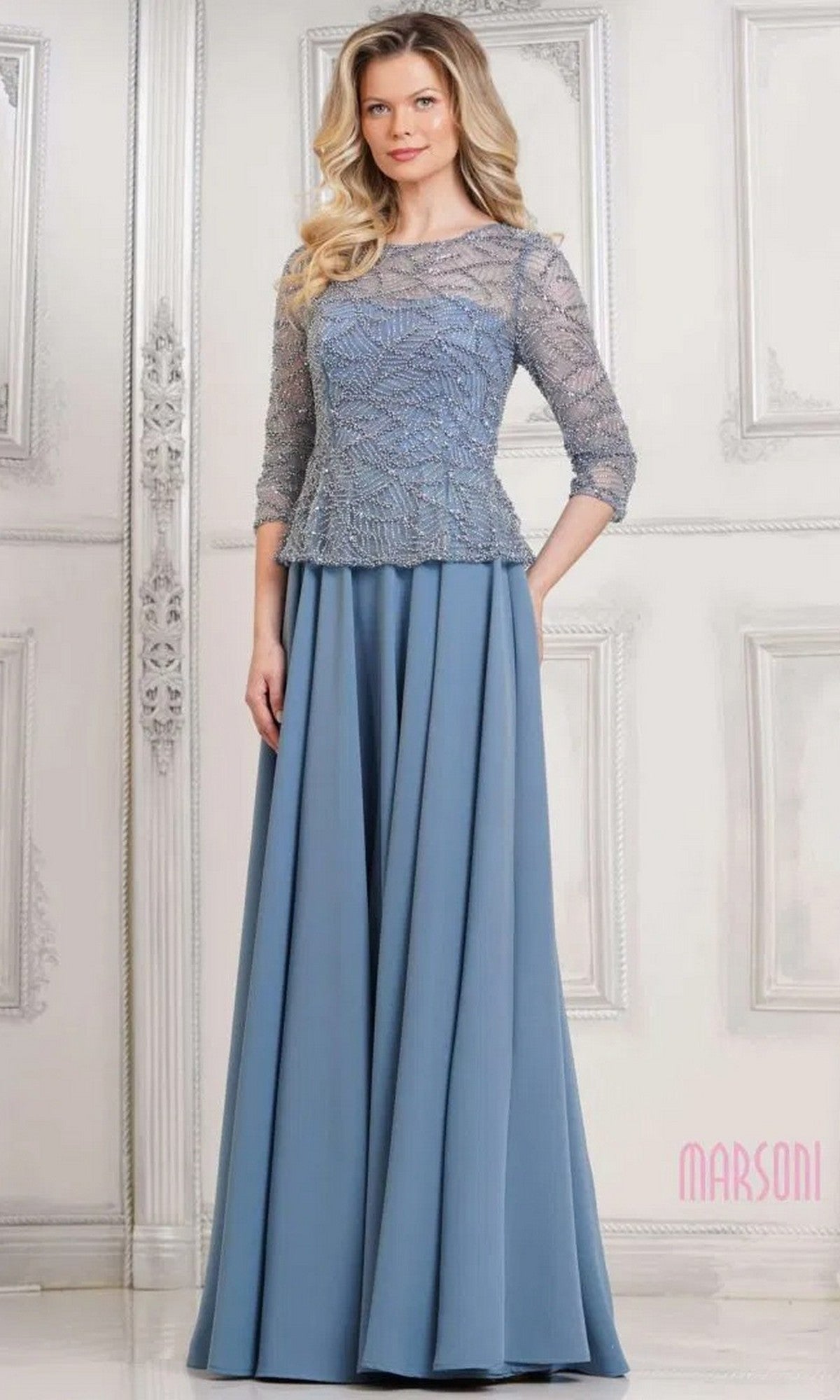 A woman in the Marsoni MV1319 long formal dress, featuring a beaded bodice, three-quarter sleeves, and an elegant slate blue A-line skirt, stands before an ornate white wall.