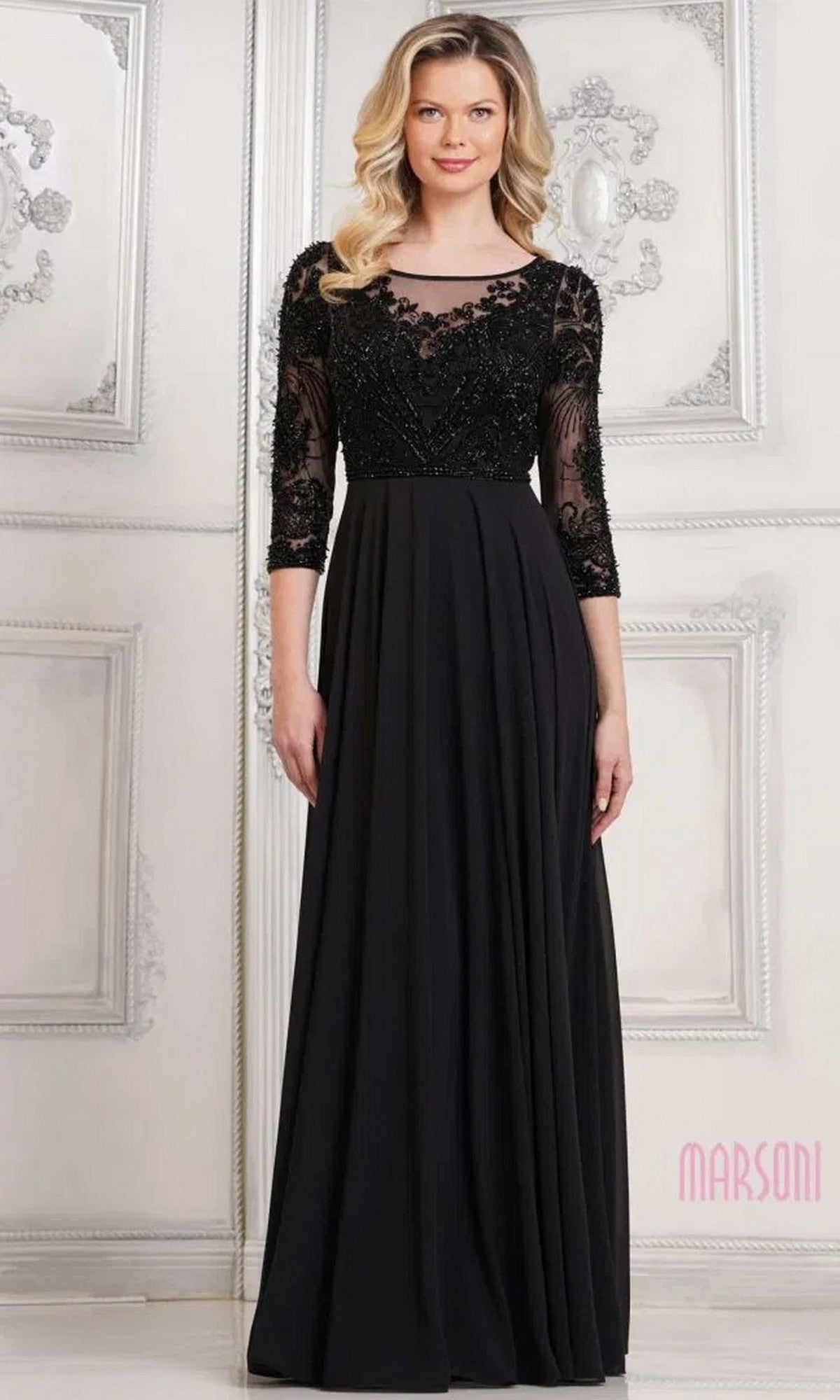 A woman in the black Marsoni MV1322 long formal dress, featuring a sequin-embellished lace bodice, three-quarter sleeves, and a flowing chiffon pleated skirt, stands in an elegantly decorated room.
