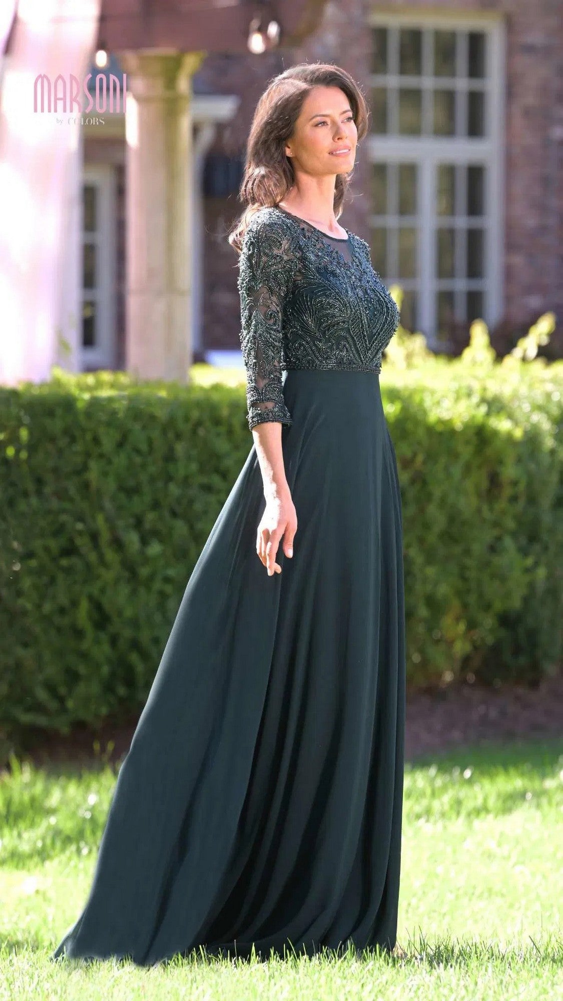A woman stands outdoors in the Marsoni MV1322 Long Formal Dress, featuring a lace embroidered bodice, fitted lace sleeves, and a flowing chiffon pleated skirt, set before green hedges and a building with large windows.