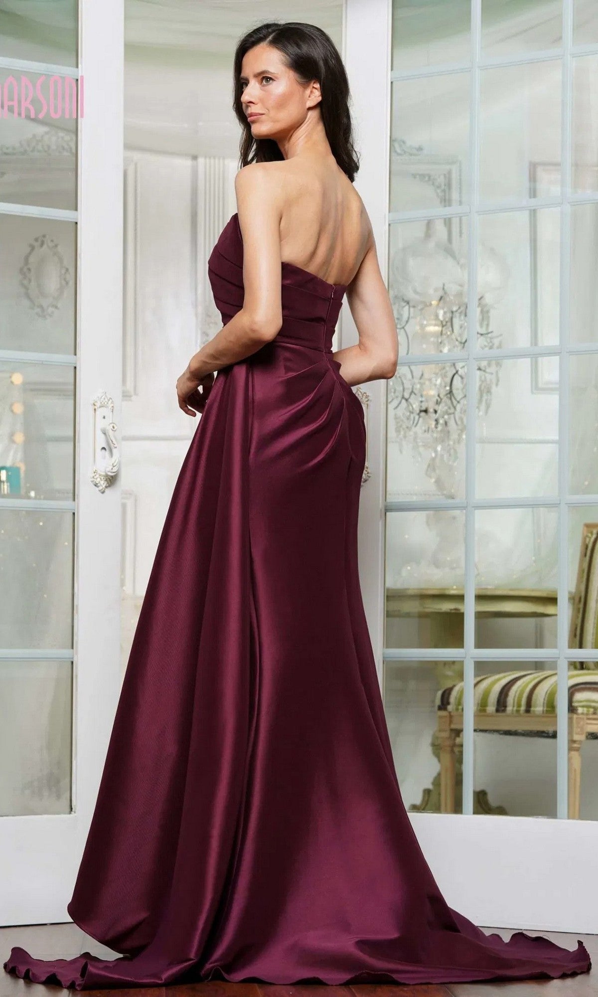 A woman poses indoors with her back partially turned, wearing the Marsoni MV1326 long formal strapless evening gown in floor-length burgundy.