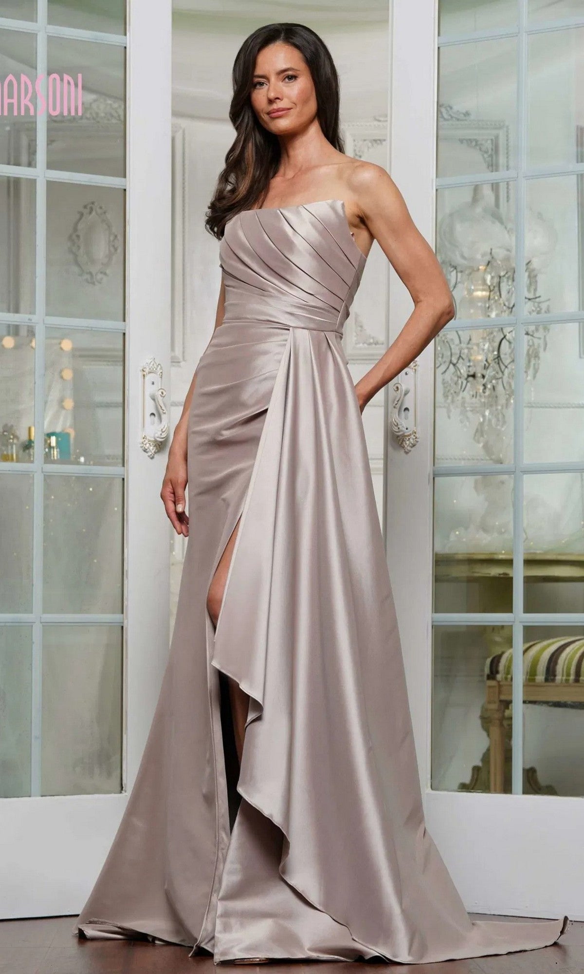 A woman stands indoors wearing the Marsoni MV1326 long formal dress in taupe, featuring a one-shoulder design, pleated detailing, and a thigh-high slit.